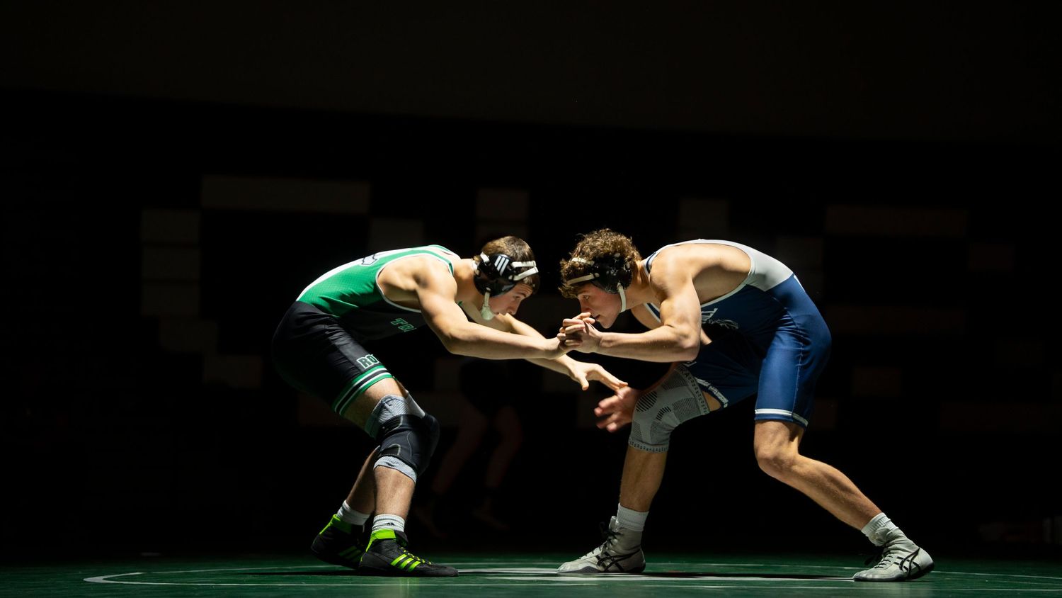 Two high school wrestlers in green and blue singlets grapple in a low stance on a wrestling mat.