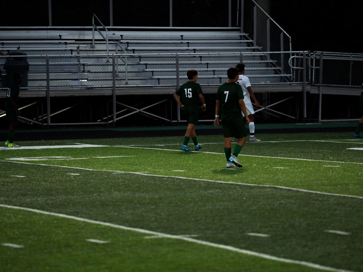 Two Trinity soccer players #15 and #7 position themselves near bleachers during nighttime match on turf.