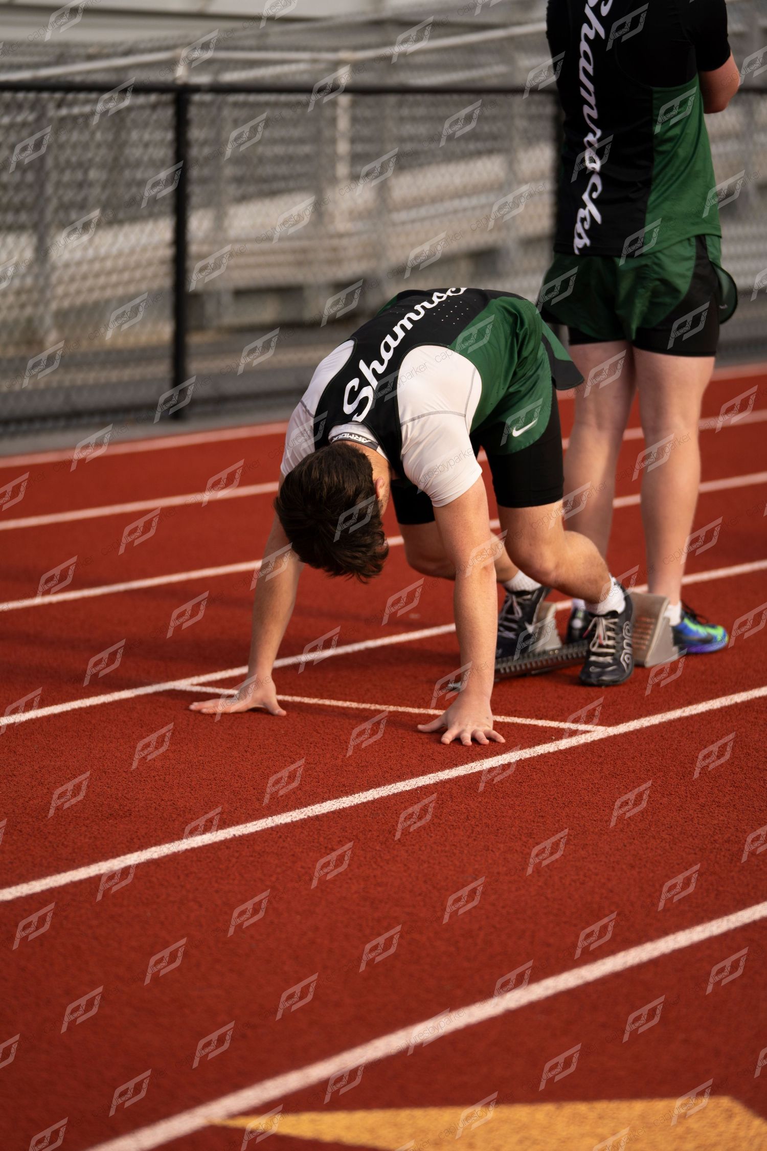 High school runner in green Shamrocks uniform crouches in starting position on red track.