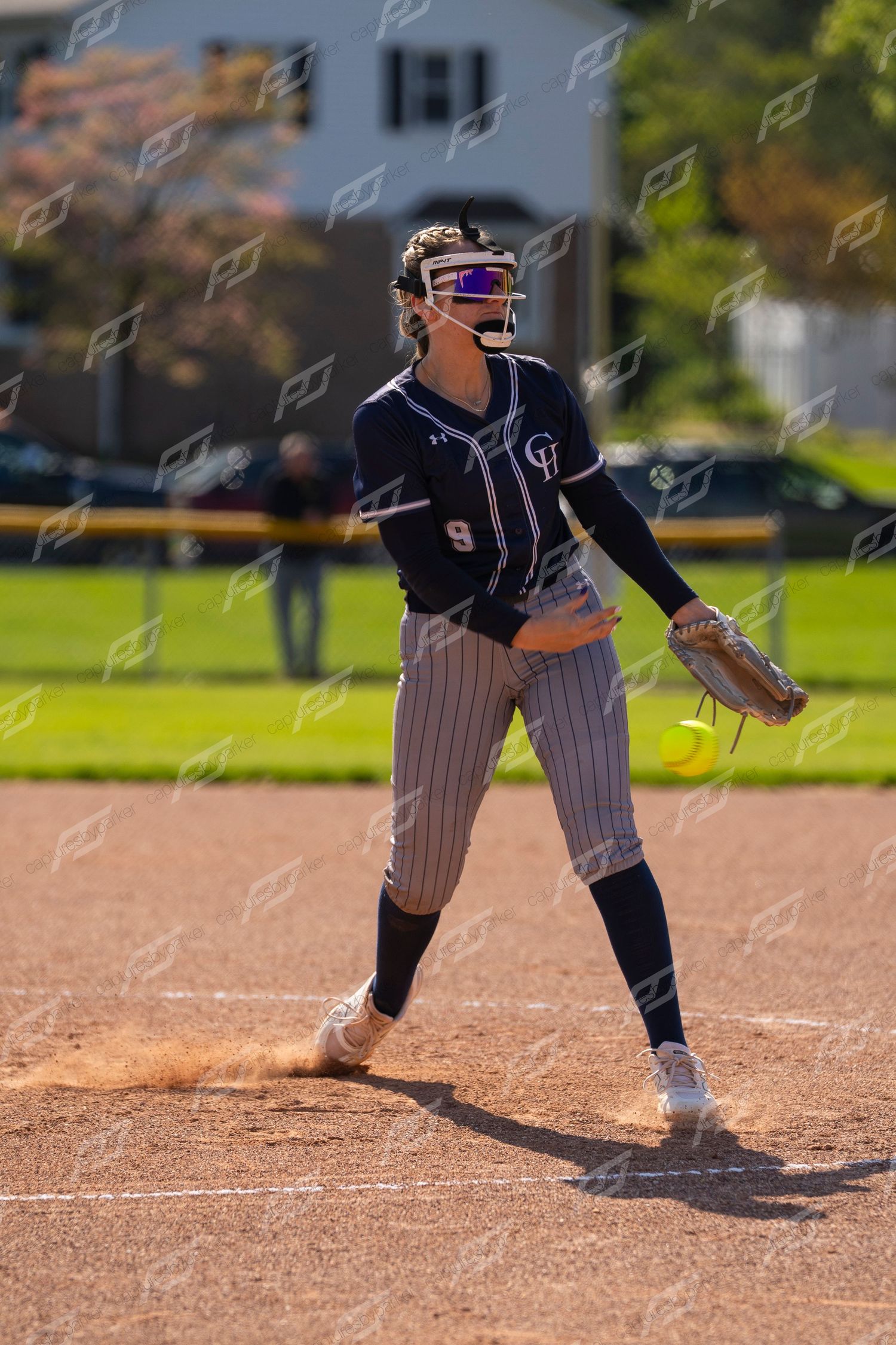 Female softball pitcher in navy pinstripe uniform #9 releases yellow ball on dirt mound.