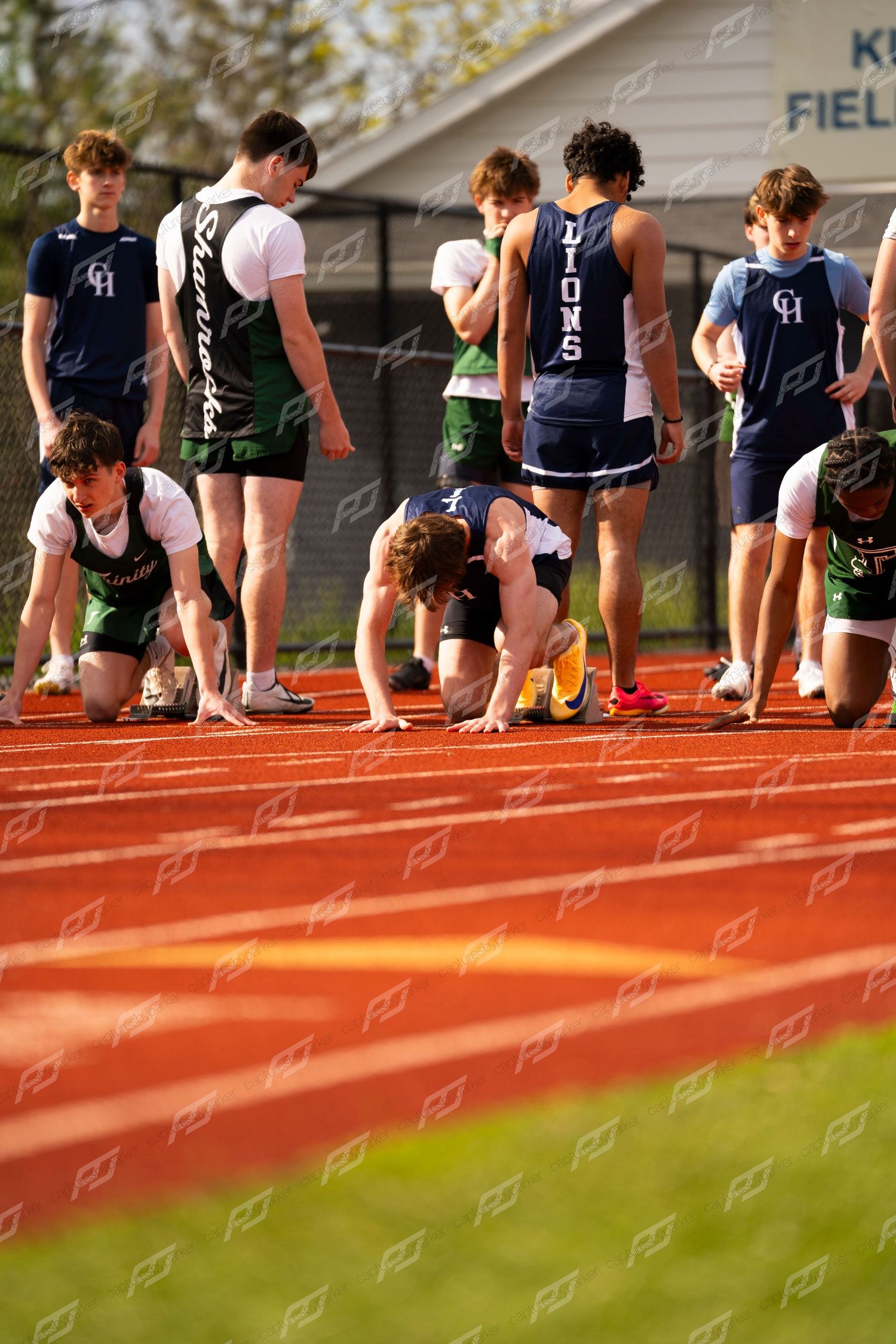 Male high school sprinters in green and navy uniforms set up at starting blocks on red track.