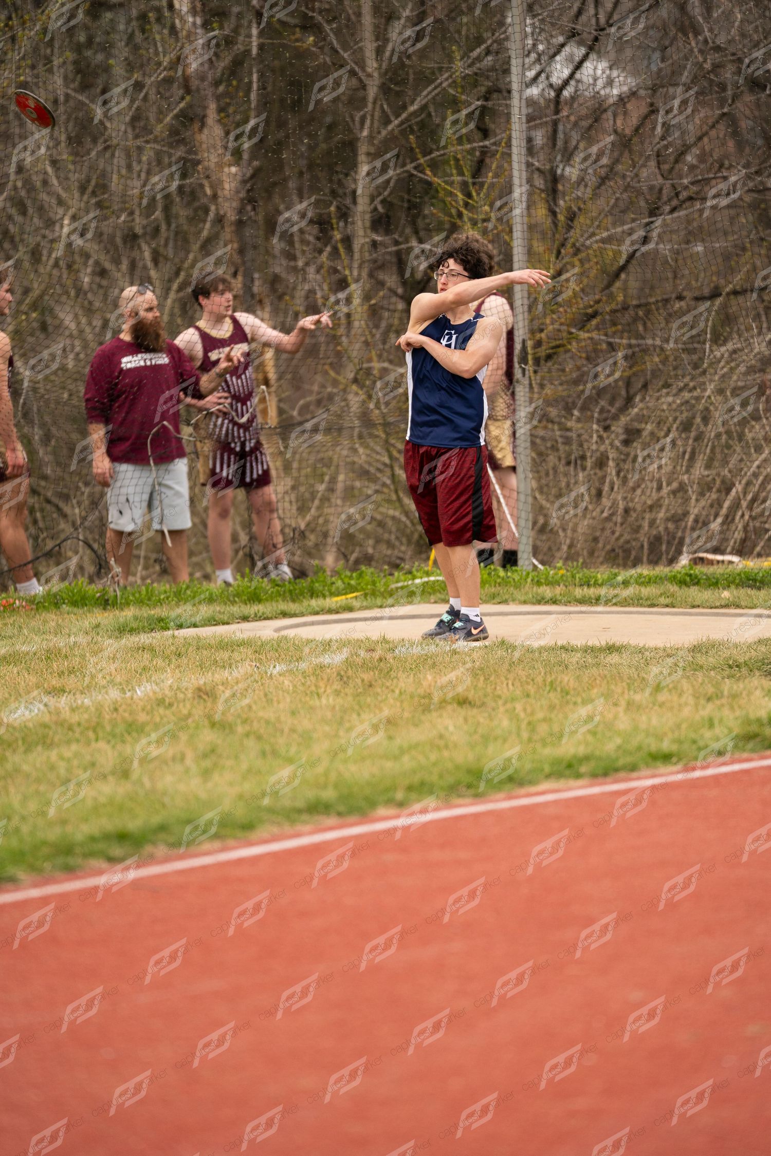 A high school athlete throws a discus during an outdoor track and field competition with spectators nearby.