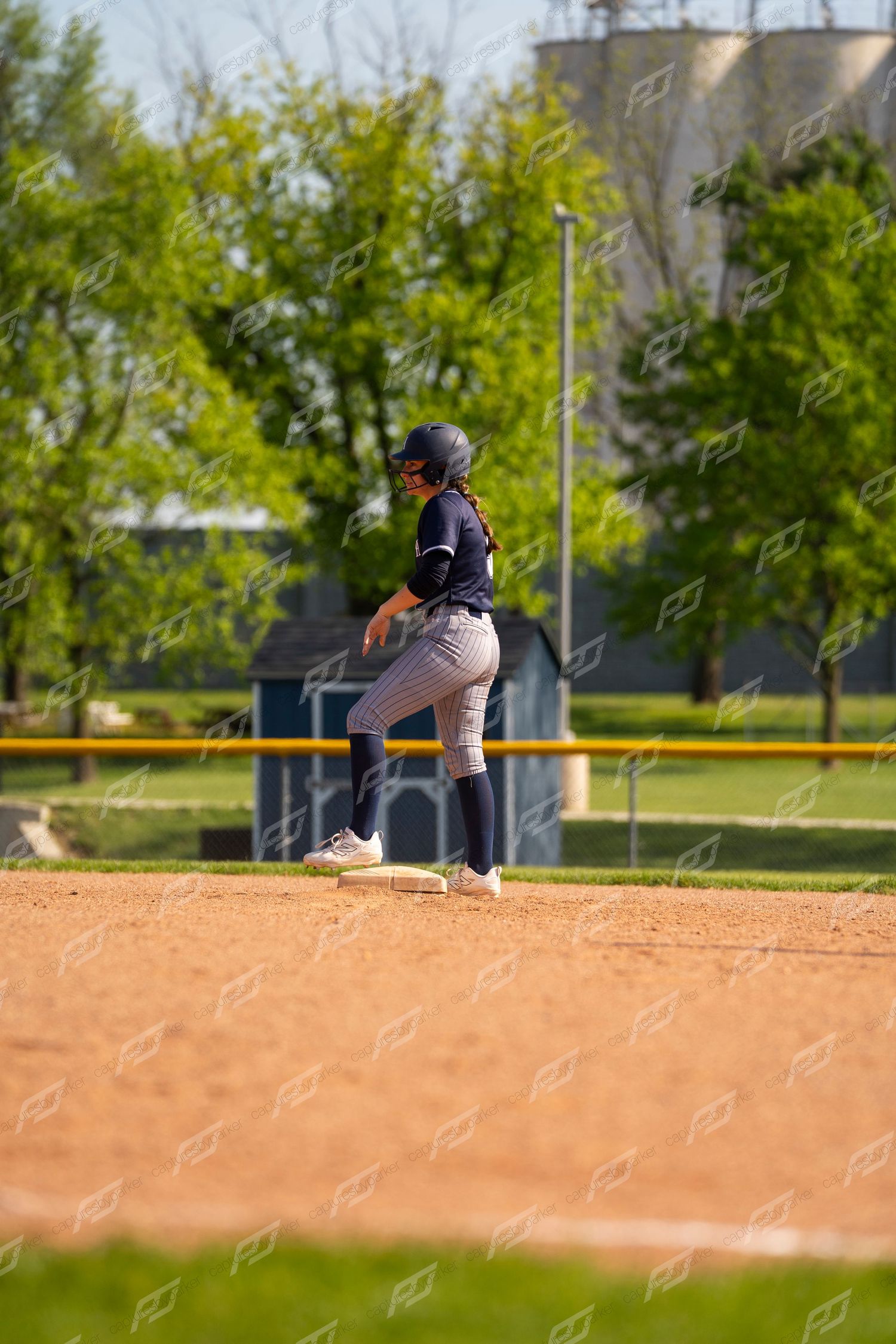 Softball player in navy uniform stands on base during a sunny outdoor game with green trees visible in background.