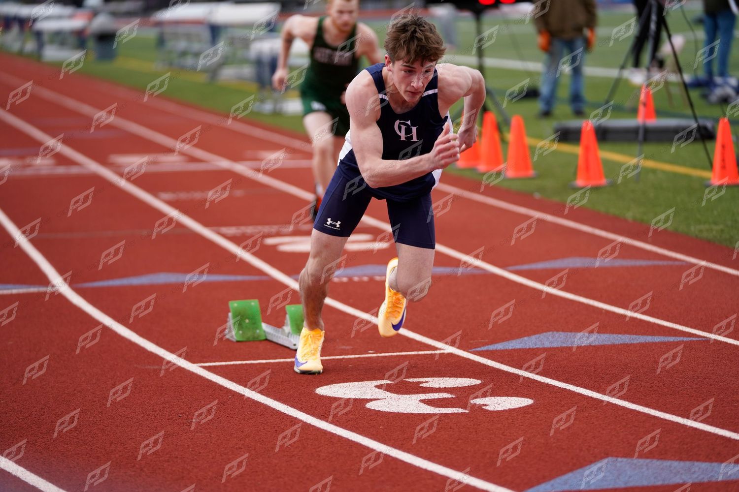 Male sprinter exploding off starting blocks on red track wearing navy uniform and yellow spikes.