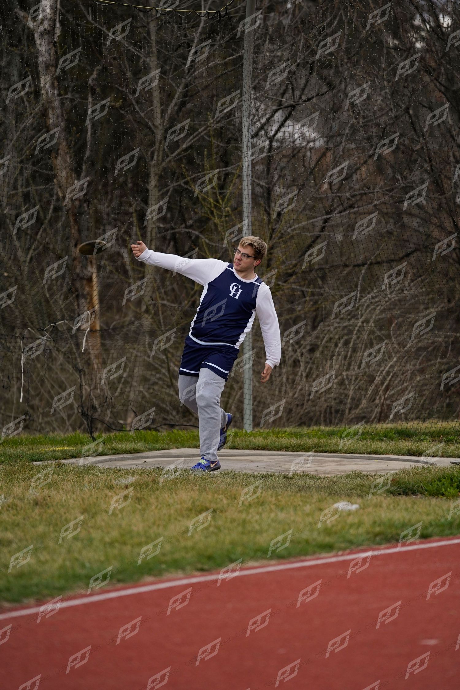 Male athlete in navy varsity jacket releasing a discus at an outdoor track and field meet.