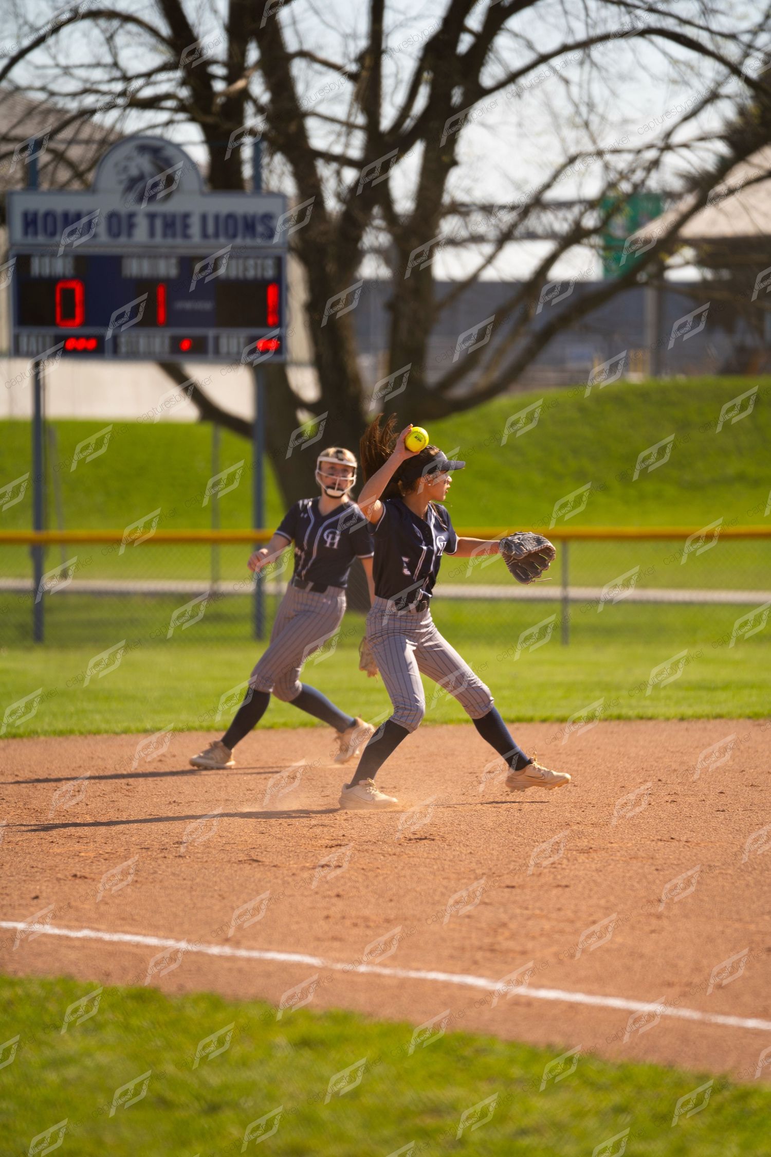 Two softball players in navy uniforms field the ball near the pitcher's mound.