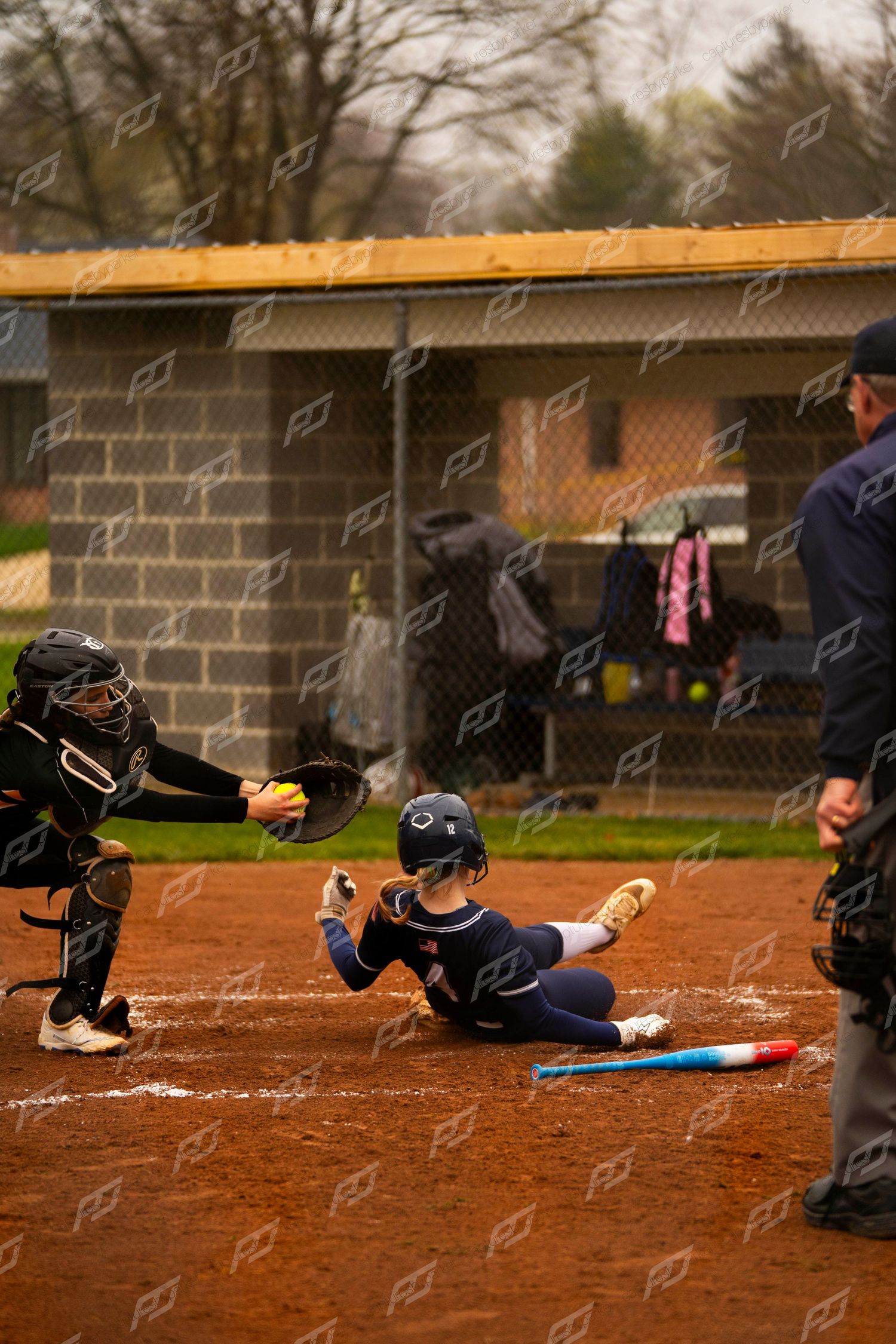 Softball player slides into home plate as catcher receives the ball with umpire watching closely.