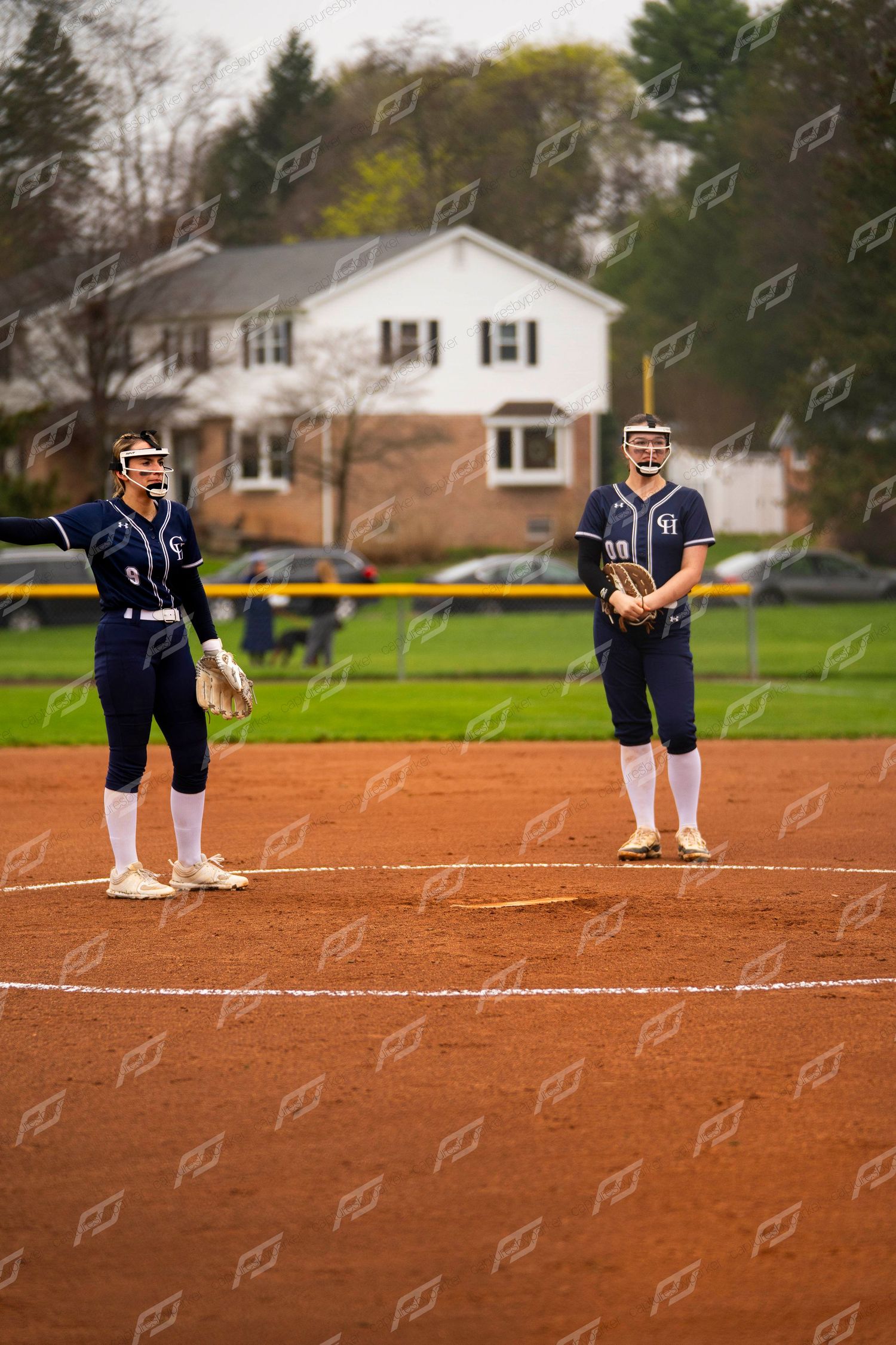 Two softball players in navy uniforms stand near first base on a red dirt field during a game.