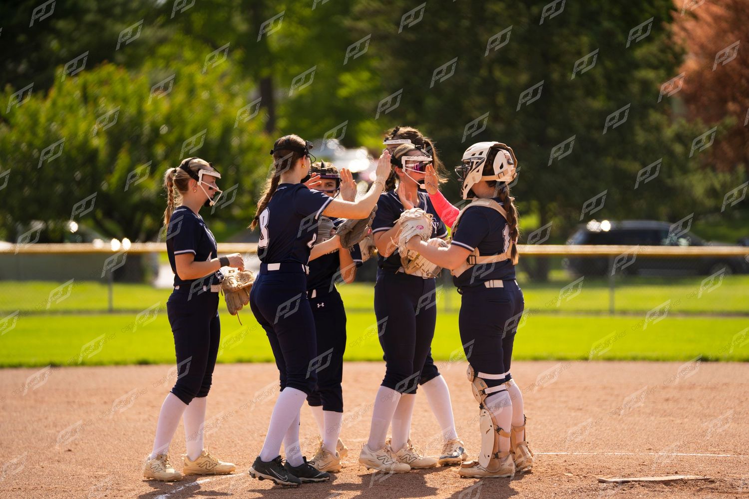 Five softball players in navy uniforms celebrate together on the field after a play.