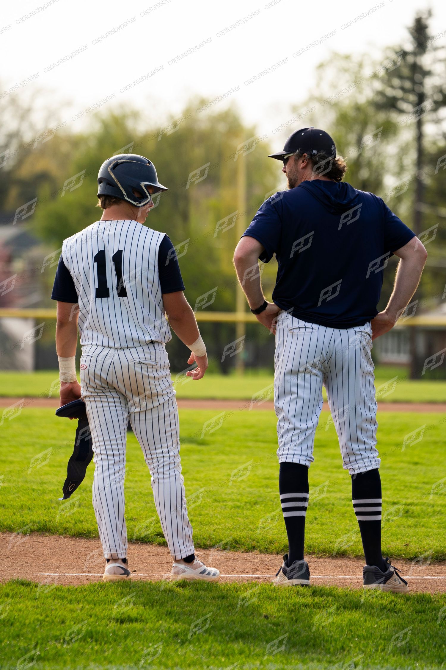 Baseball player number 11 in white pinstripes stands at third base alongside a coach during a game.