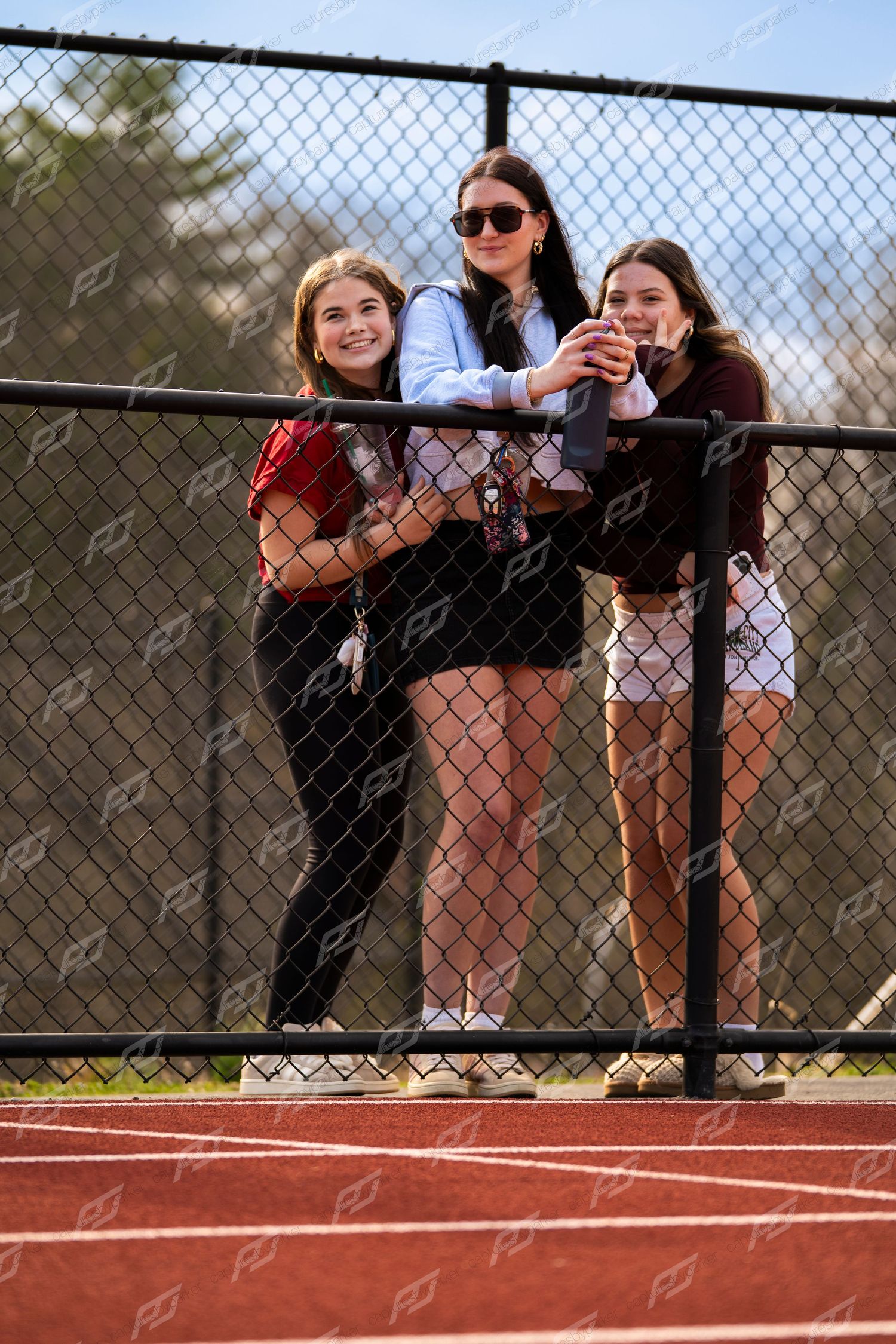 Three teenage girls smiling and posing by chain-link fence at outdoor track meet.