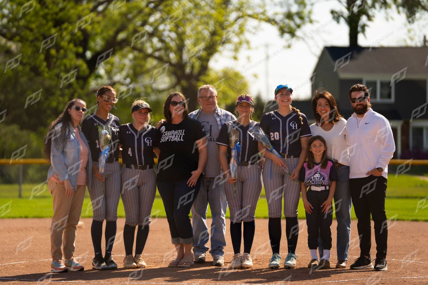 Group photo of softball team in navy uniforms with families gathered together on sunny baseball field.