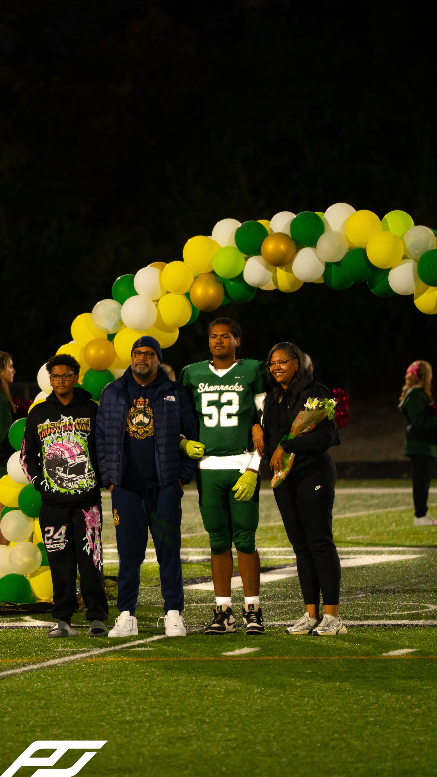 Shamrocks football player #52 poses with family under balloon arch during senior night on green turf field.