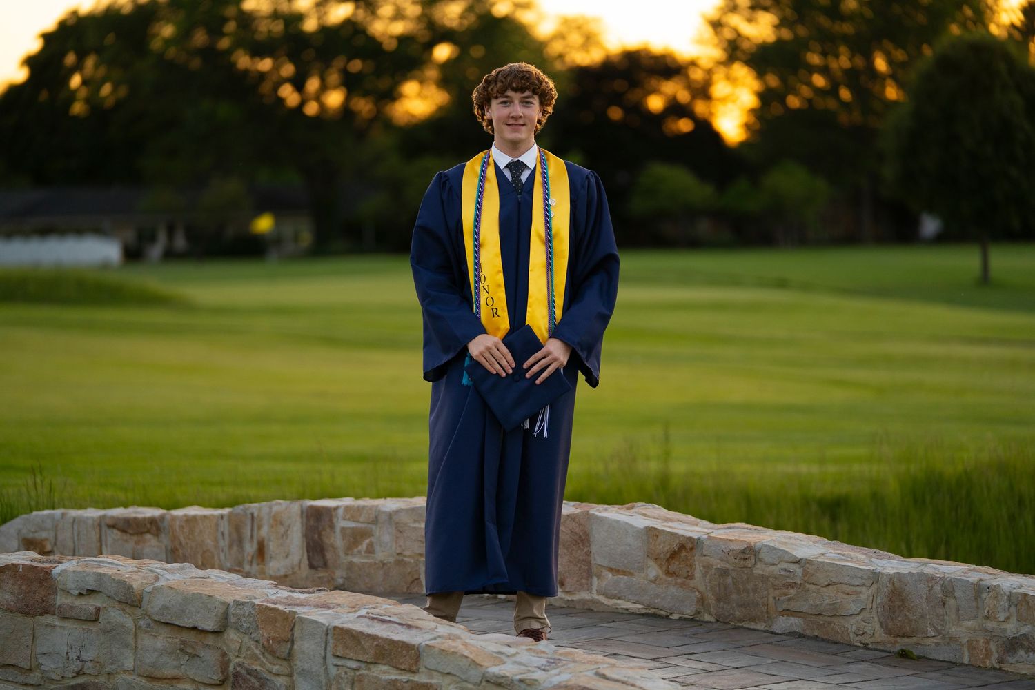 Young graduate poses in navy blue cap and gown with gold stole at sunset on stone bridge.