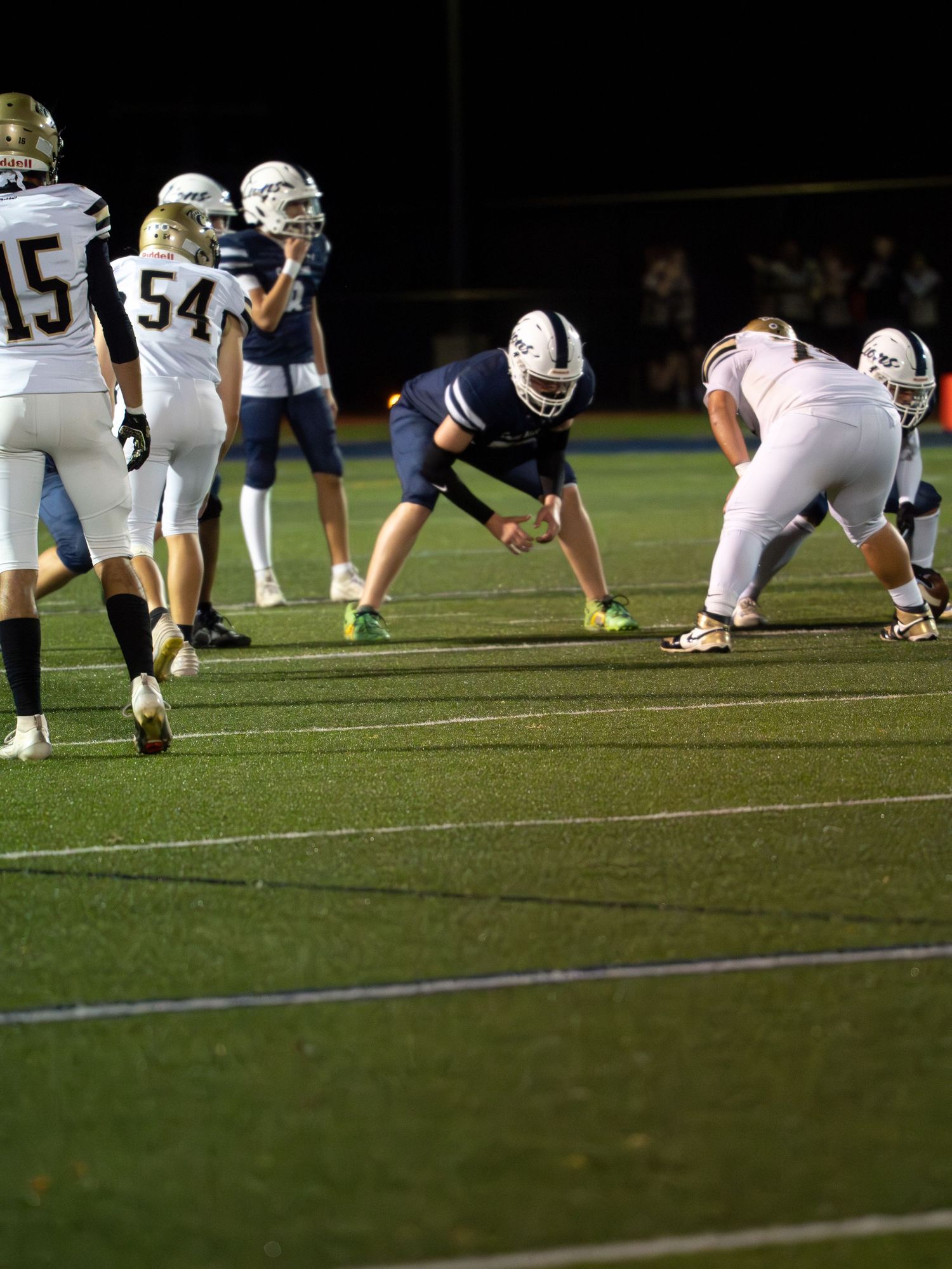 Camp Hill Lions football player in defensive stance facing opponents in gold uniforms during night game.