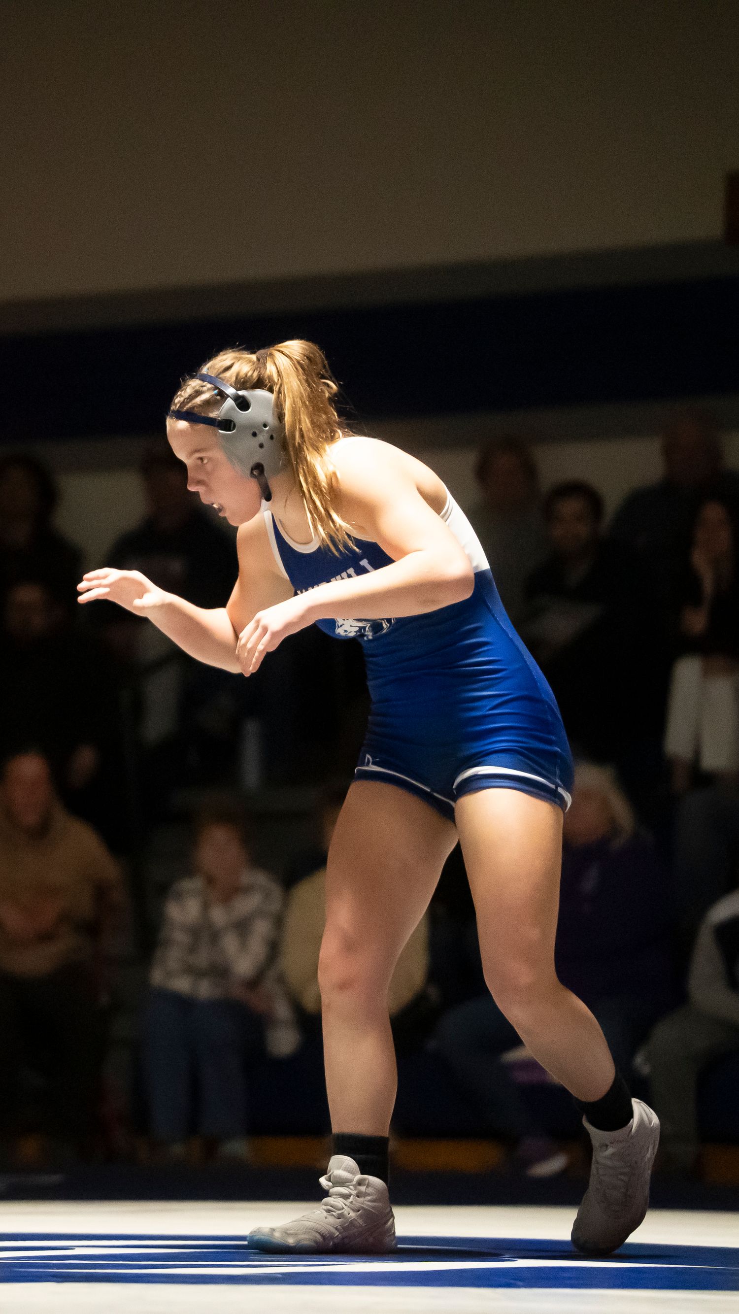 Female high school wrestler in blue singlet and headgear stands ready on the wrestling mat.