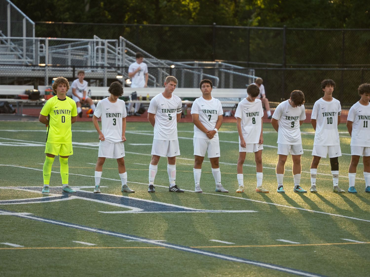Trinity soccer team lined up on field before match in white uniforms with green goalkeeper jersey.