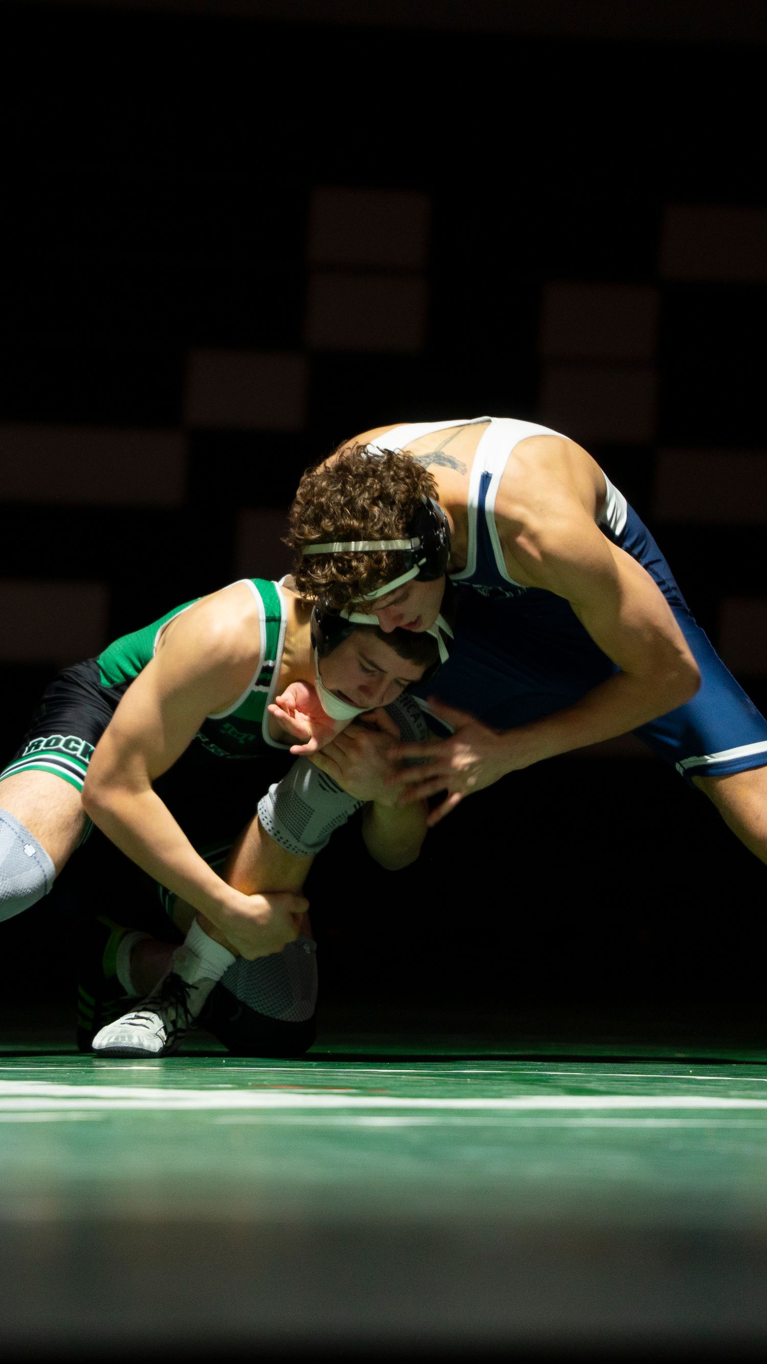 Close-up of two wrestlers competing, one in blue and one in green singlet, locked in a tight hold.