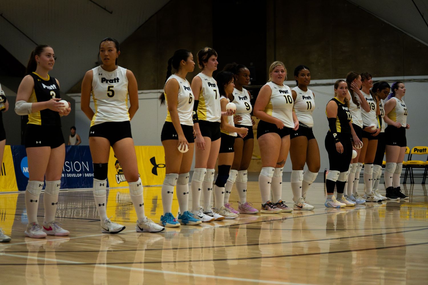 Women's volleyball team in black and gold uniforms lined up on indoor court during pre-game ceremony.