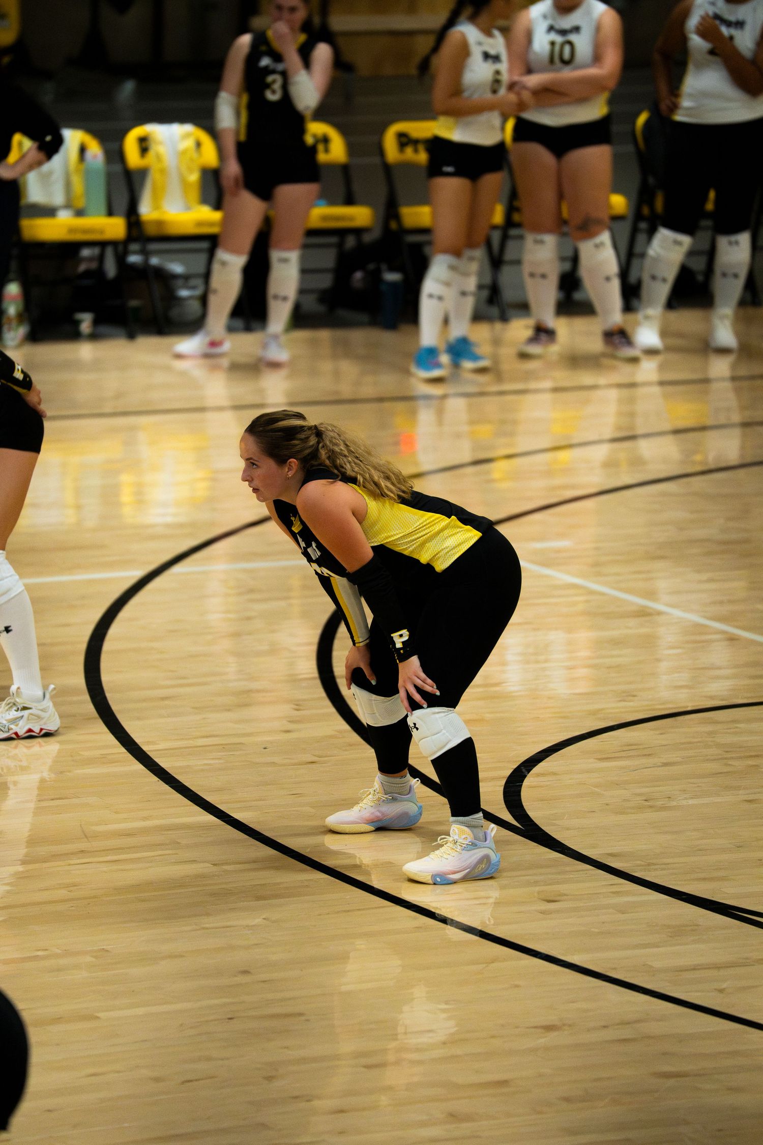 Female volleyball player in yellow and black uniform crouches in ready position on indoor court during a match.
