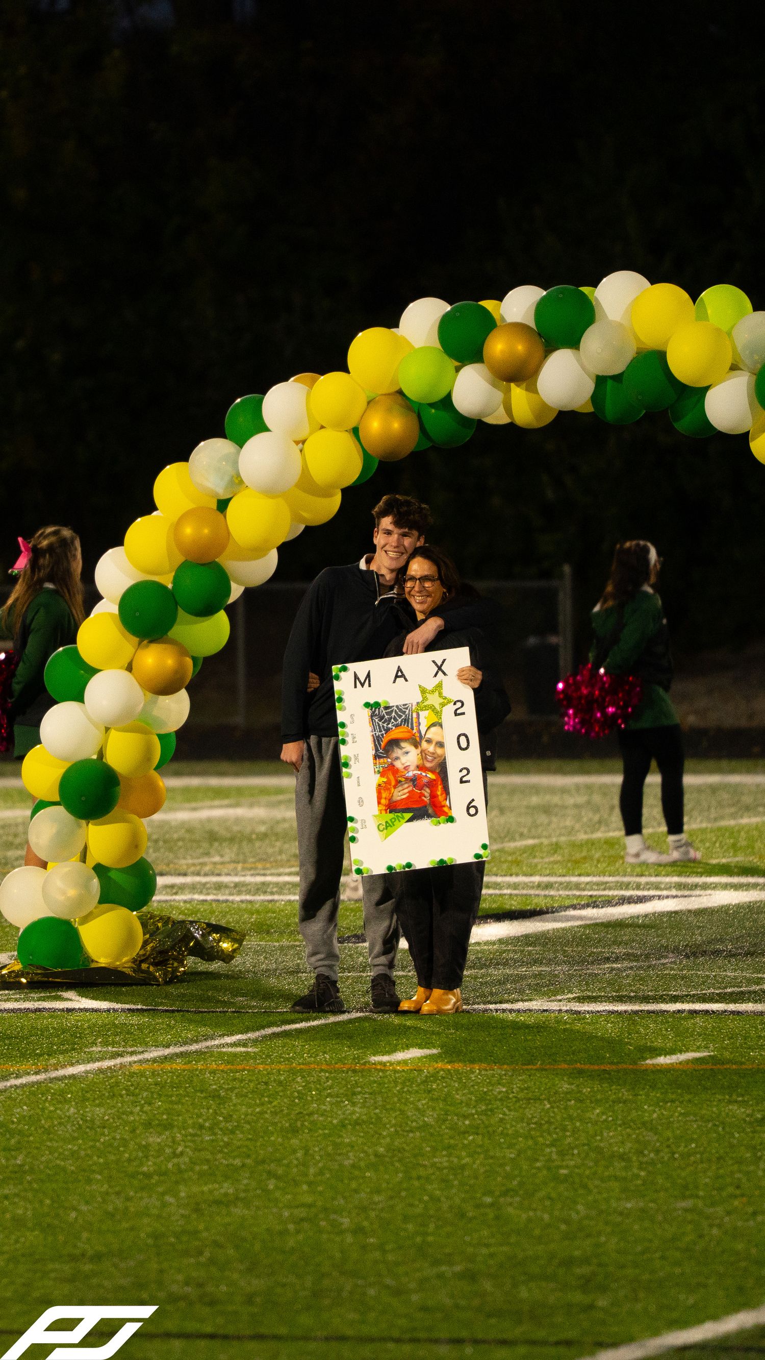 Max 2026 senior night celebration with companion under festive green and gold balloon arch on turf field.
