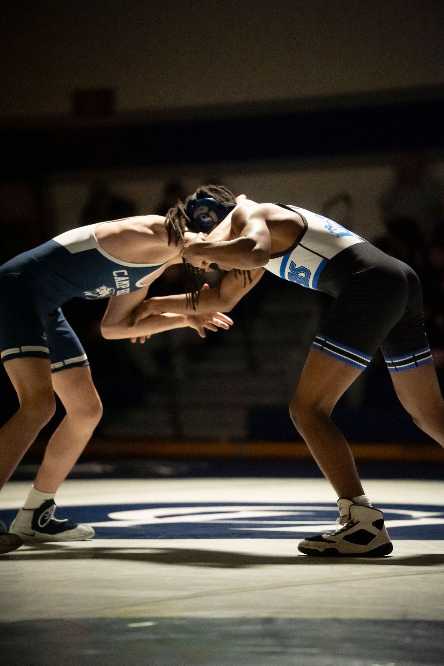 Two wrestlers in blue and white singlets lock up in a tie-up position on a dimly lit wrestling mat.