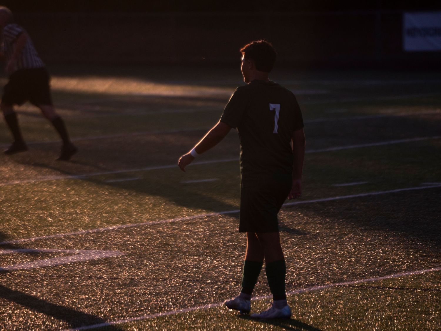 Soccer player number 7 in dark uniform walks across a sunlit turf field during an evening match.