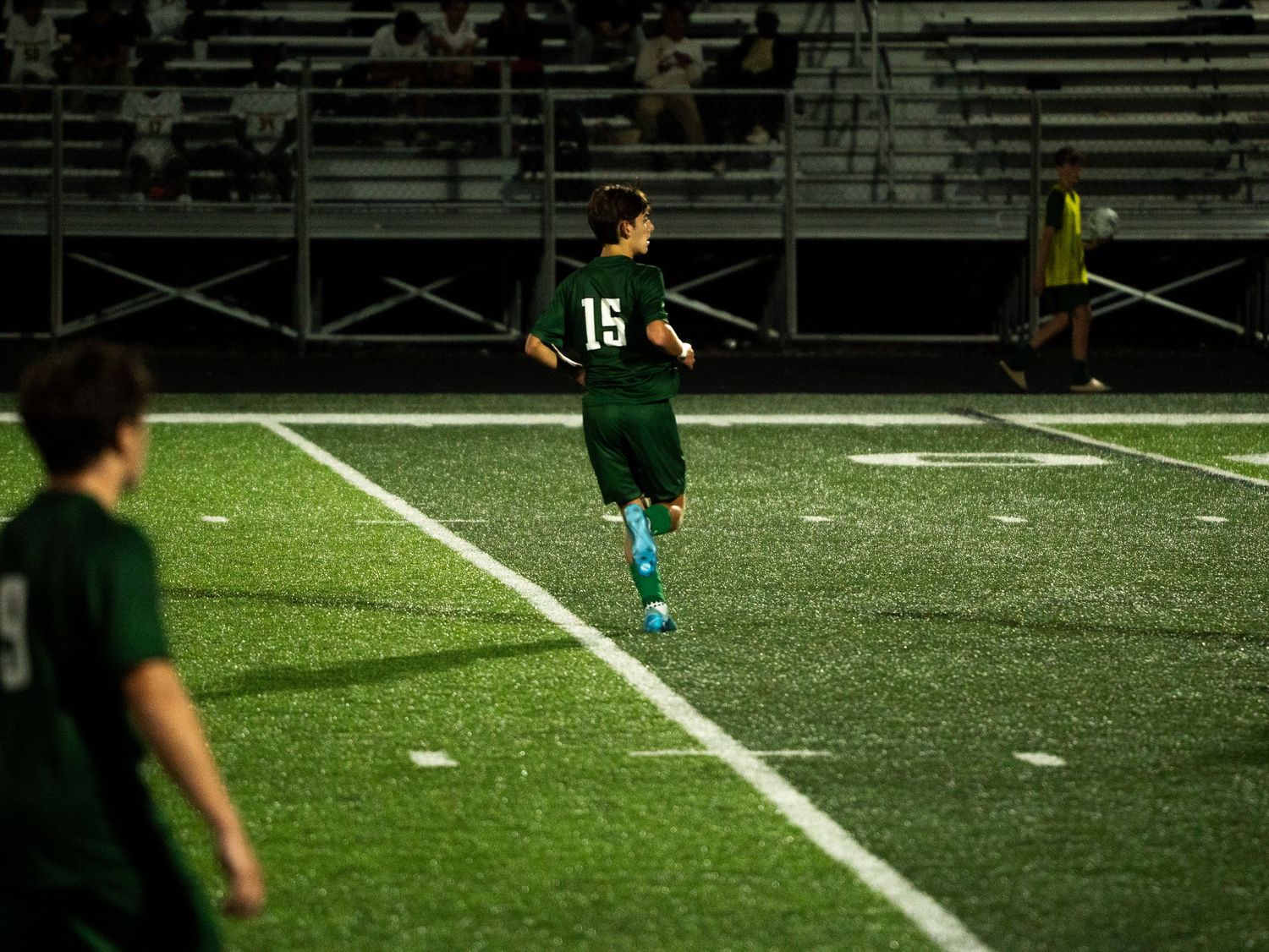 Youth soccer player wearing green number 15 jersey runs on illuminated artificial turf at night.