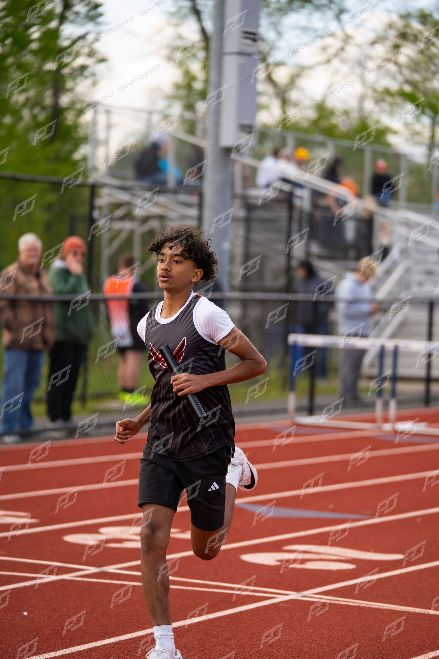 Young male track athlete in black uniform running a relay race while holding a baton on a red outdoor track.