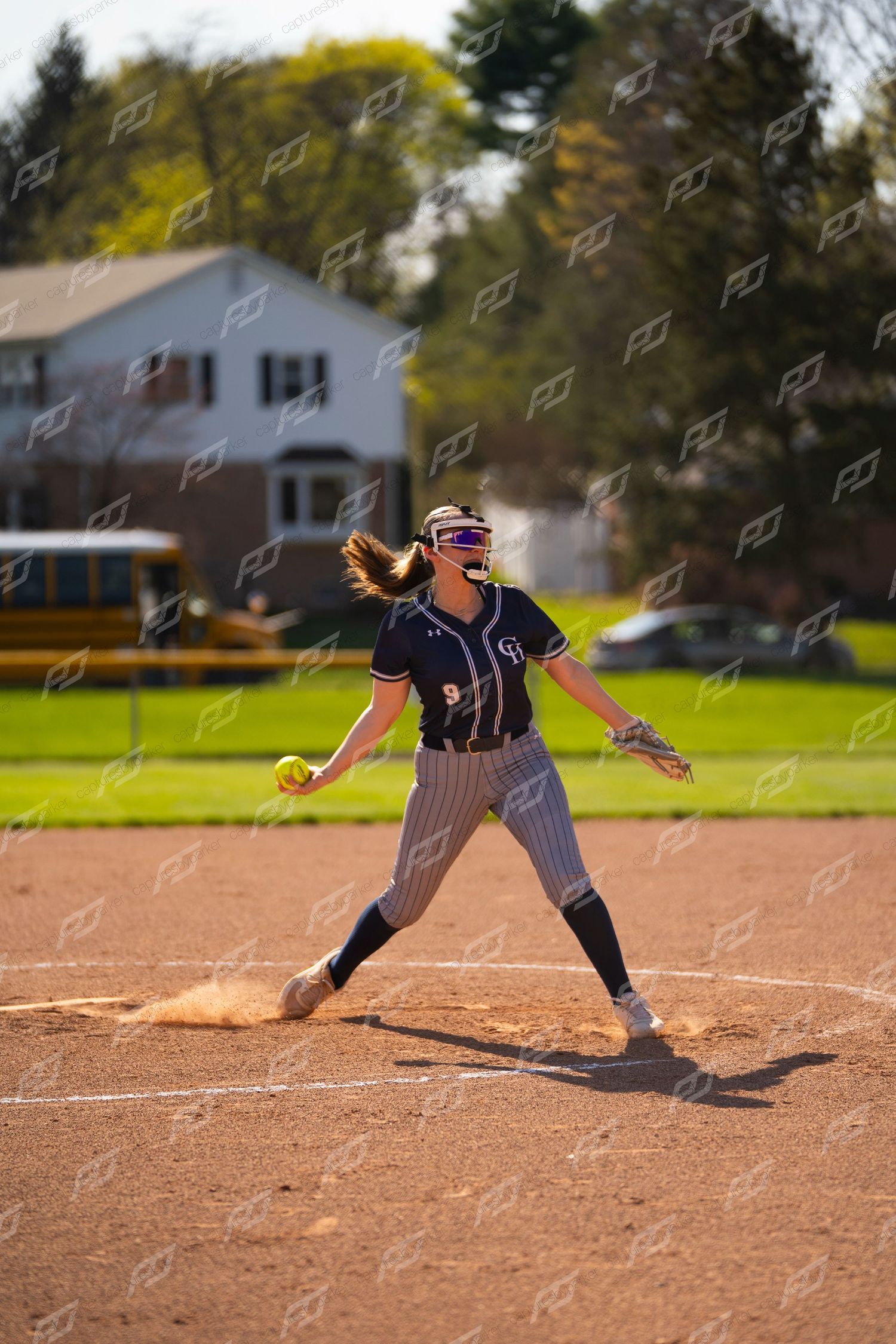 Female softball pitcher in navy pinstripe uniform delivers a pitch during an outdoor game.