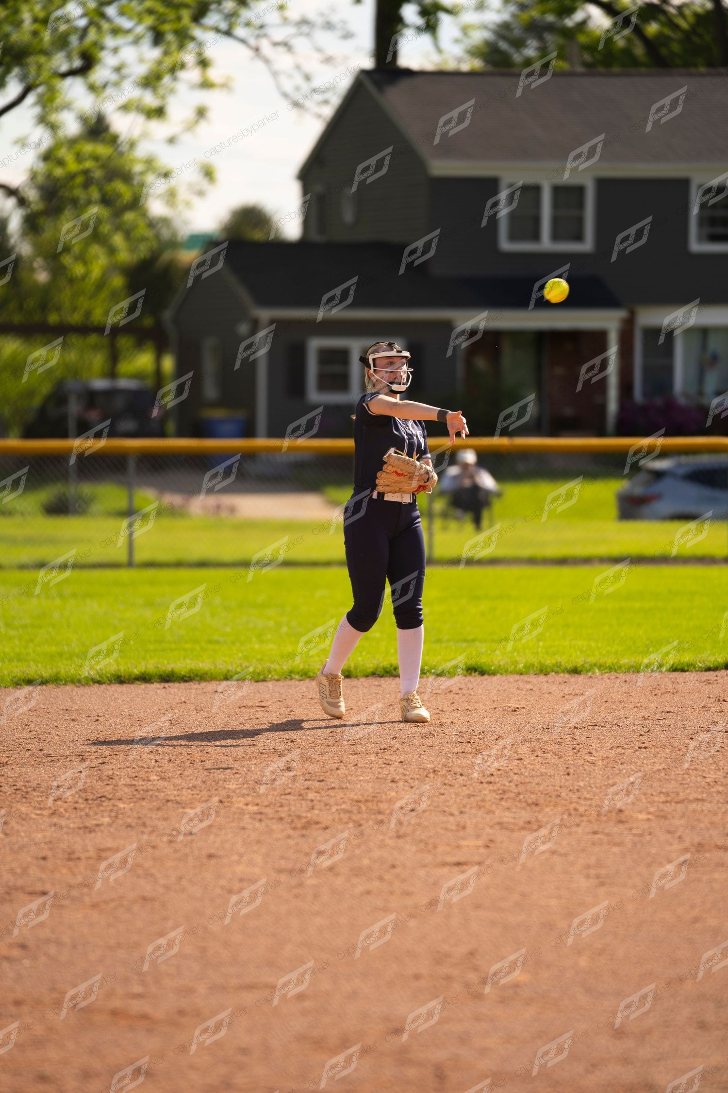 Female softball player in black uniform throws ball across infield dirt during outdoor game.