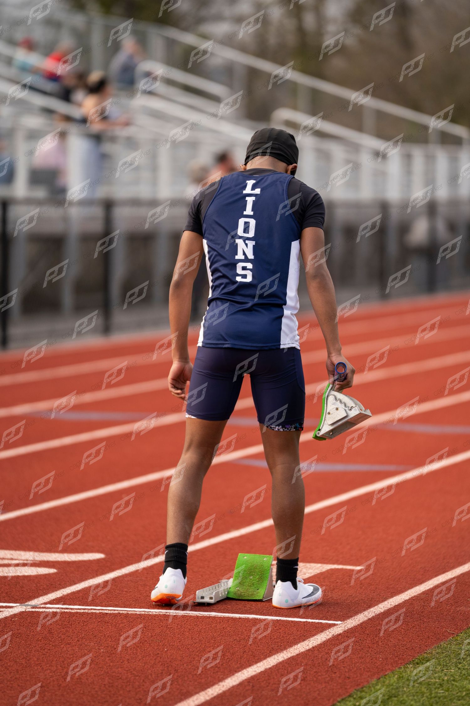 Lions track athlete stands on red track holding starting blocks, viewed from behind in navy uniform.