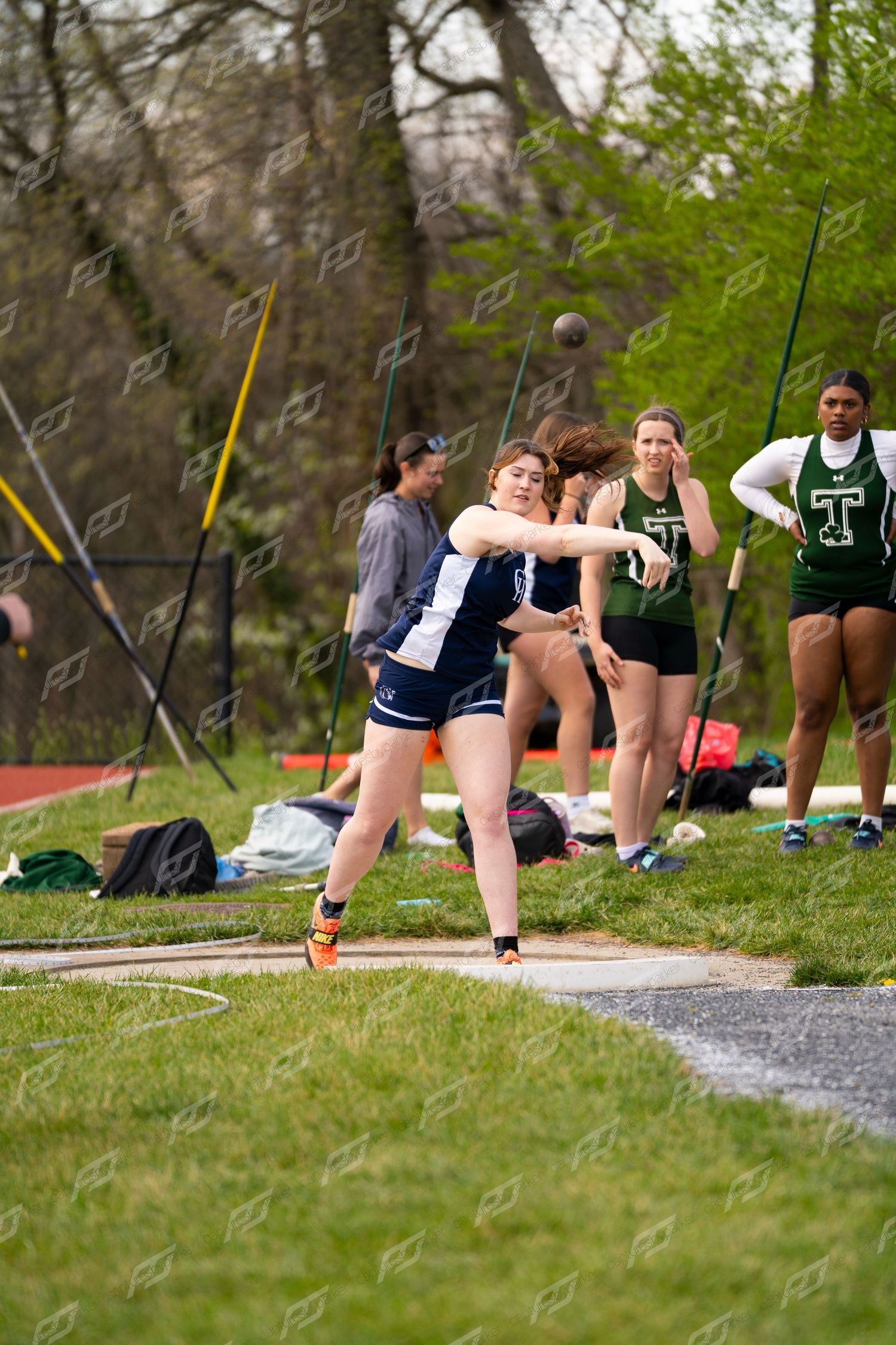 Female athlete in navy uniform releases a shot put while competitors watch in the background.