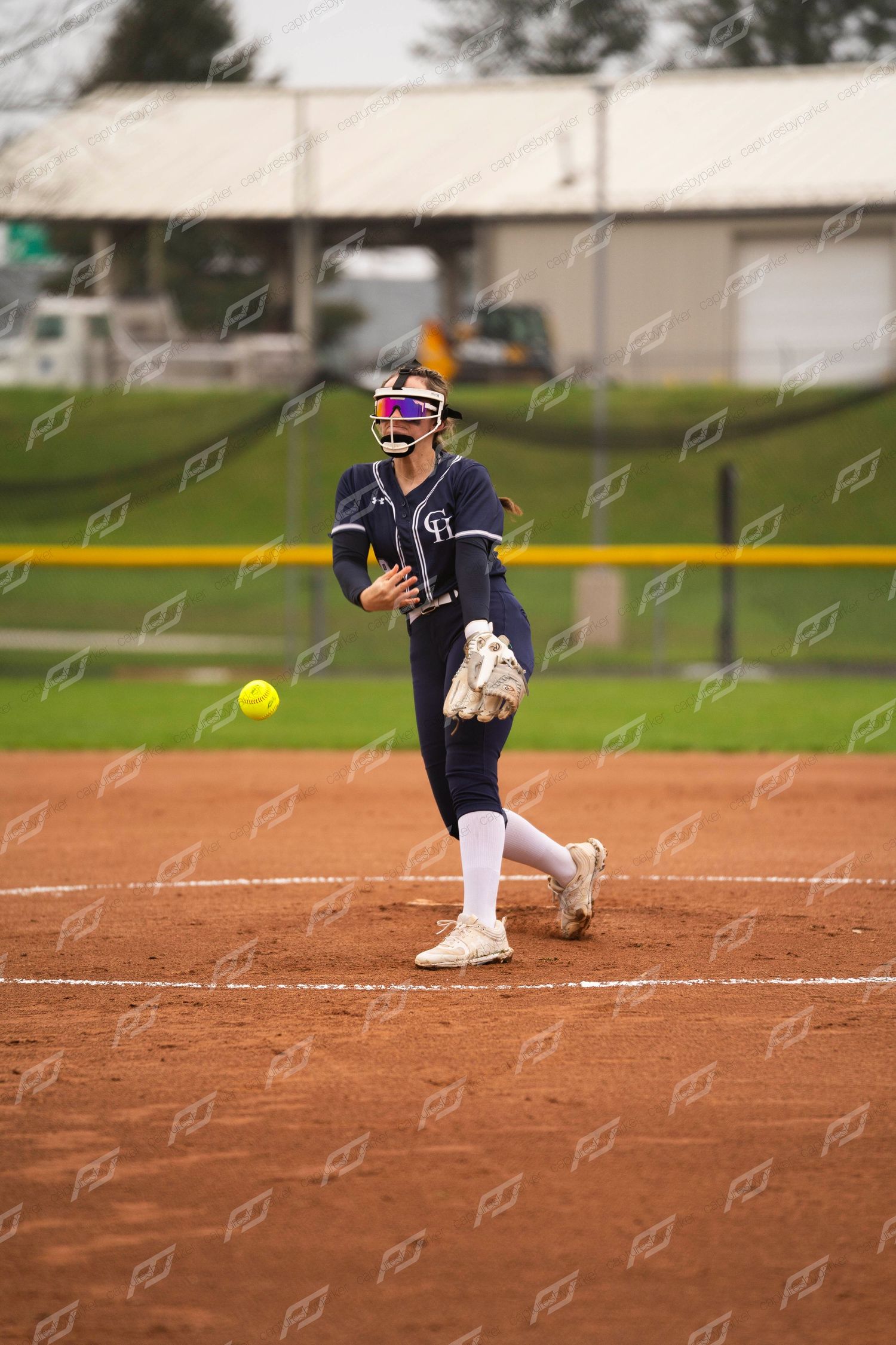 Softball pitcher in navy uniform with face guard releasing yellow ball during underhand delivery.
