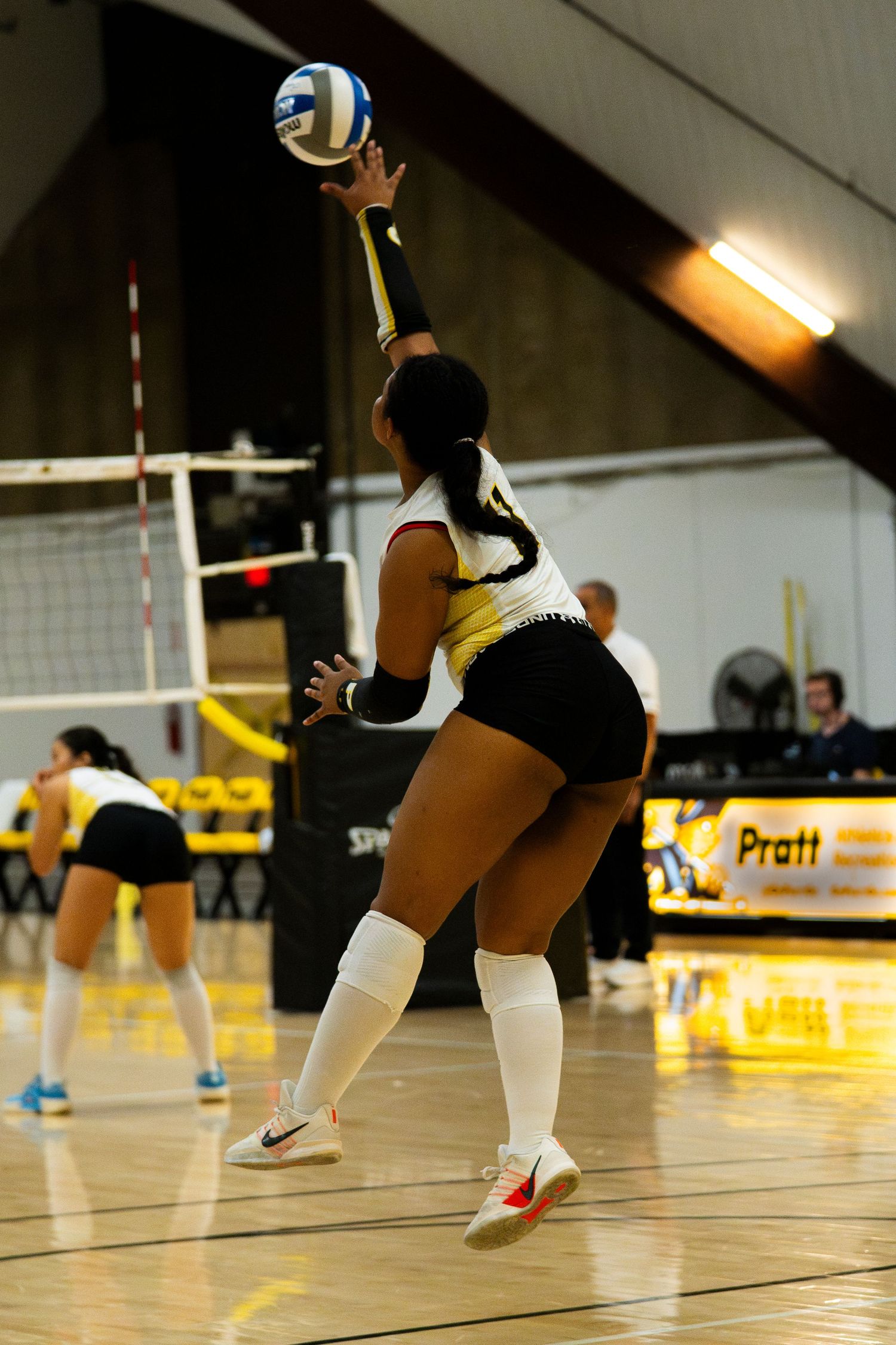 Female volleyball player in white and black uniform serving the ball during an indoor match near the net.