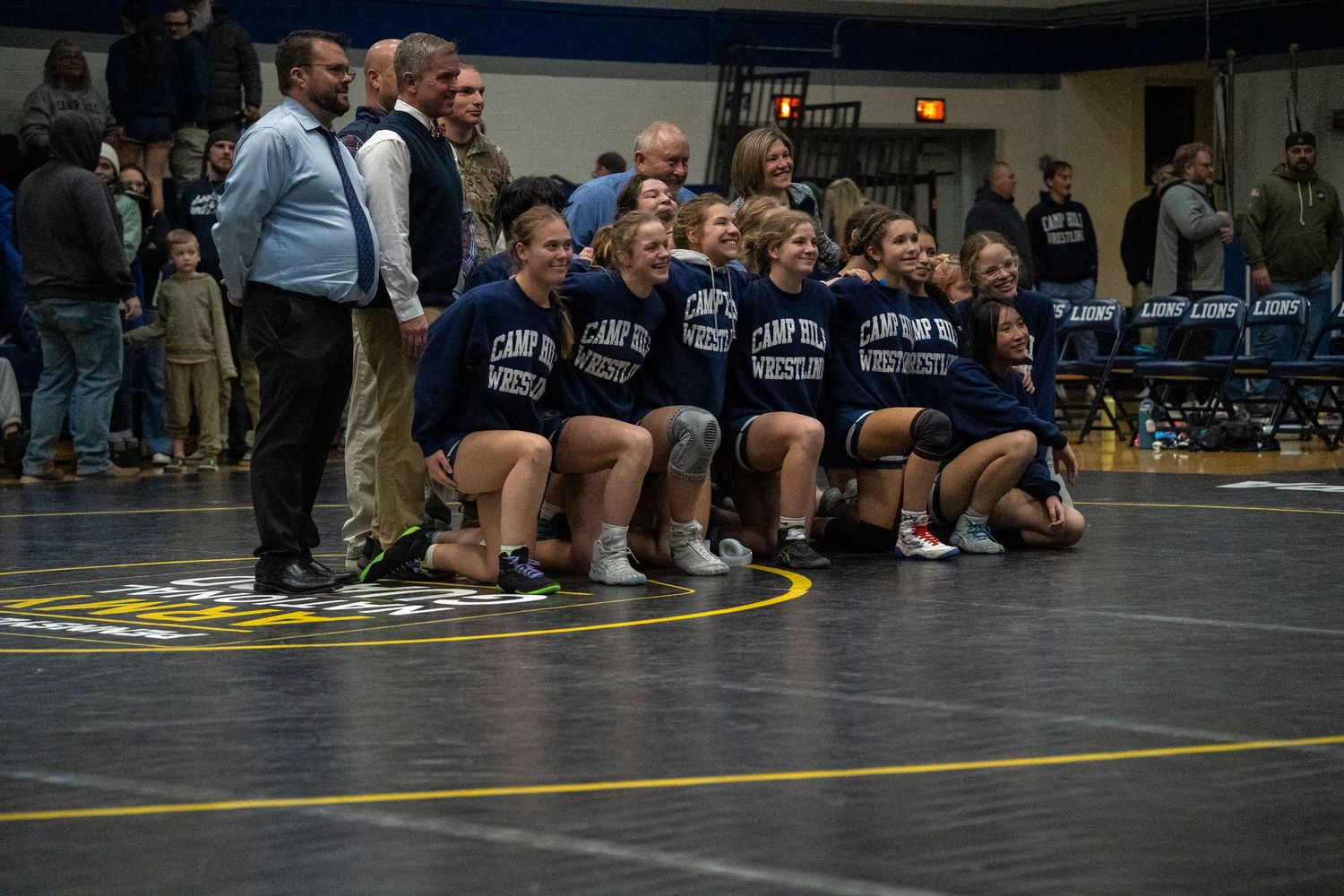 A girls wrestling team poses together on the mat smiling, wearing matching navy Camp Hill Wrestling t-shirts.