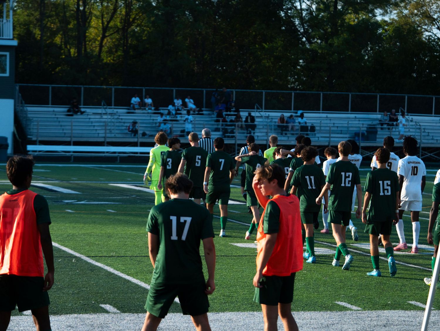 Green and white soccer teams shake hands on a turf field after a competitive high school soccer match.