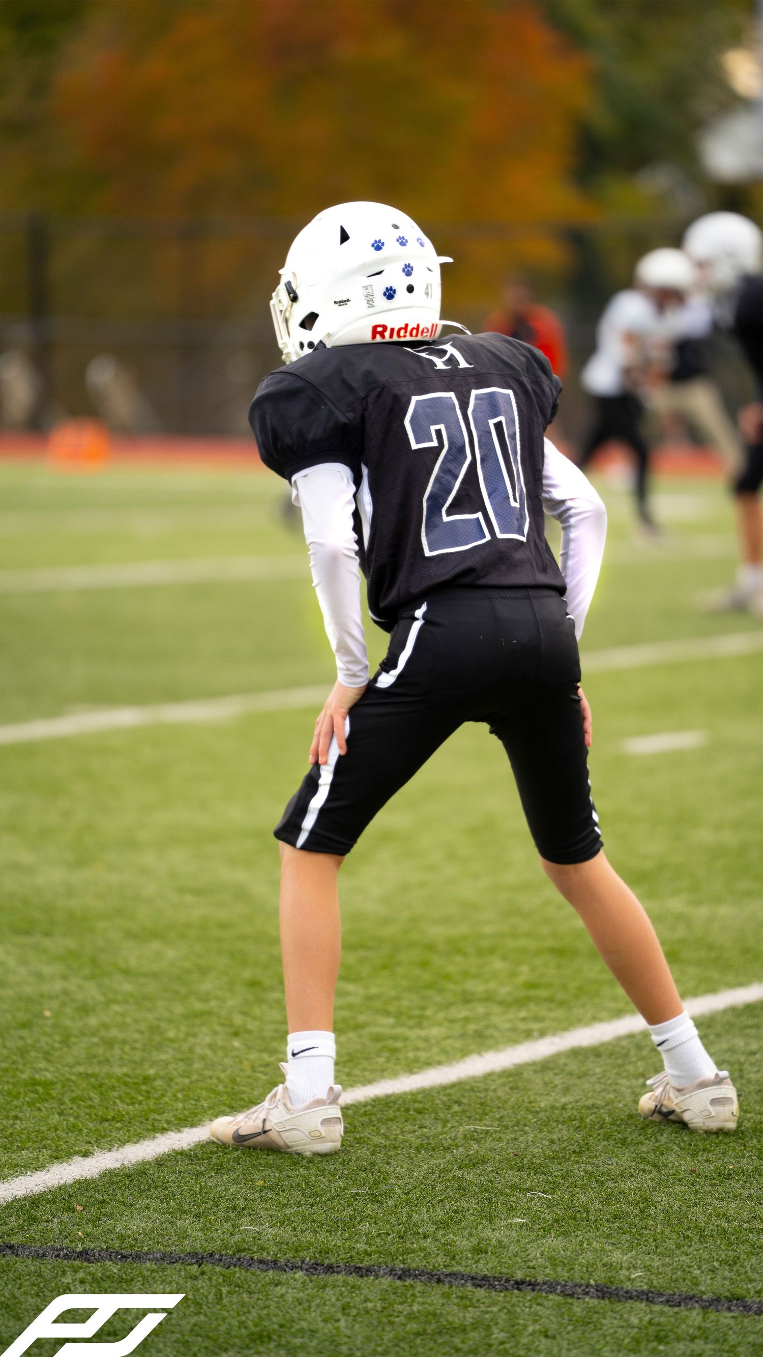 Youth football player number 20 in black uniform stands in ready position on green turf during a fall game.