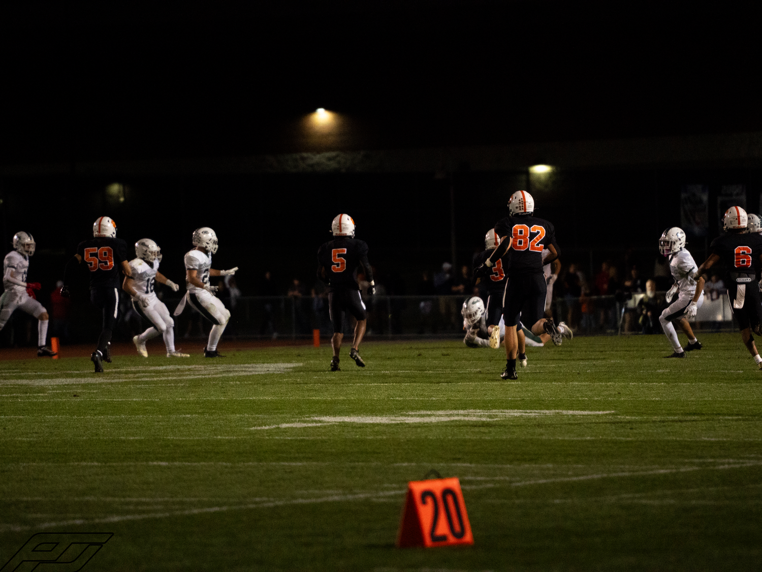 High school football players in orange and white uniforms run plays on a night game field near yard marker 20.