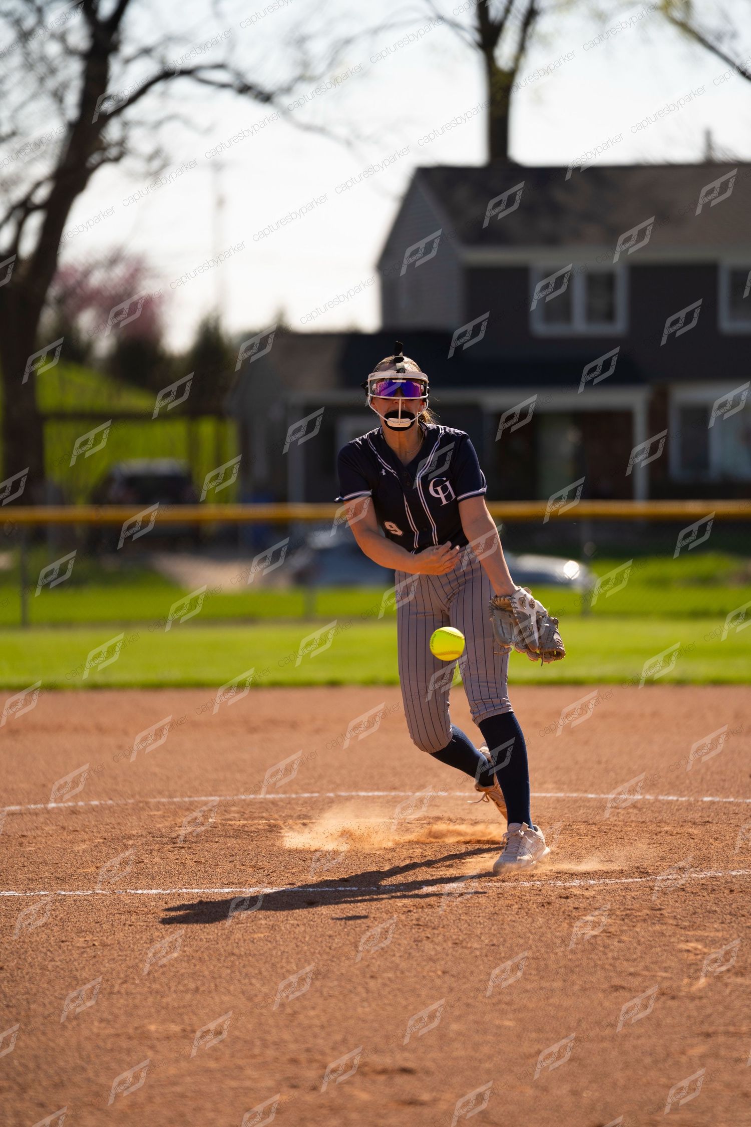 Softball pitcher in dark uniform winds up to deliver a yellow ball during evening game.