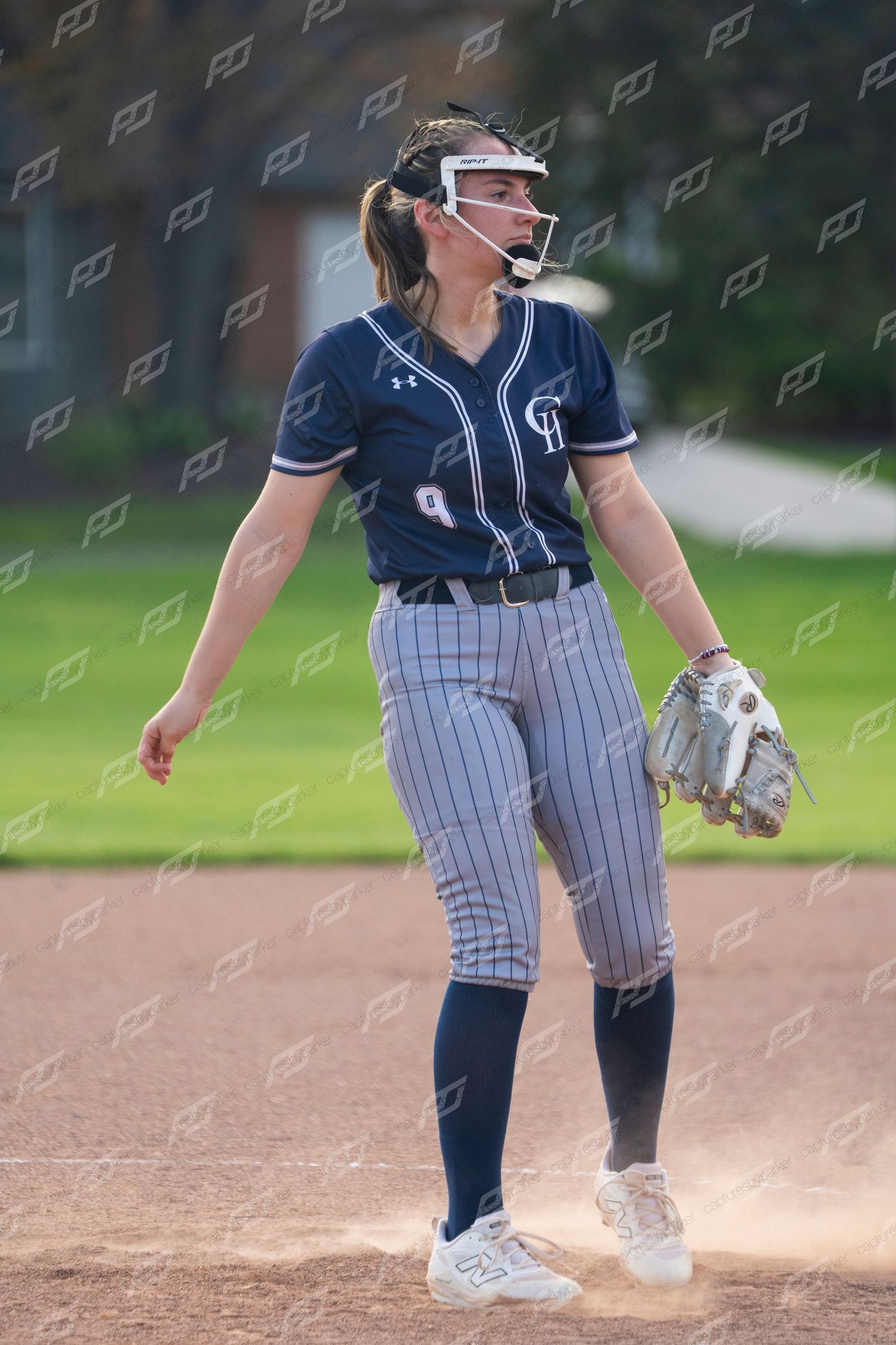 Female softball player in navy uniform with face guard stands on field holding glove.