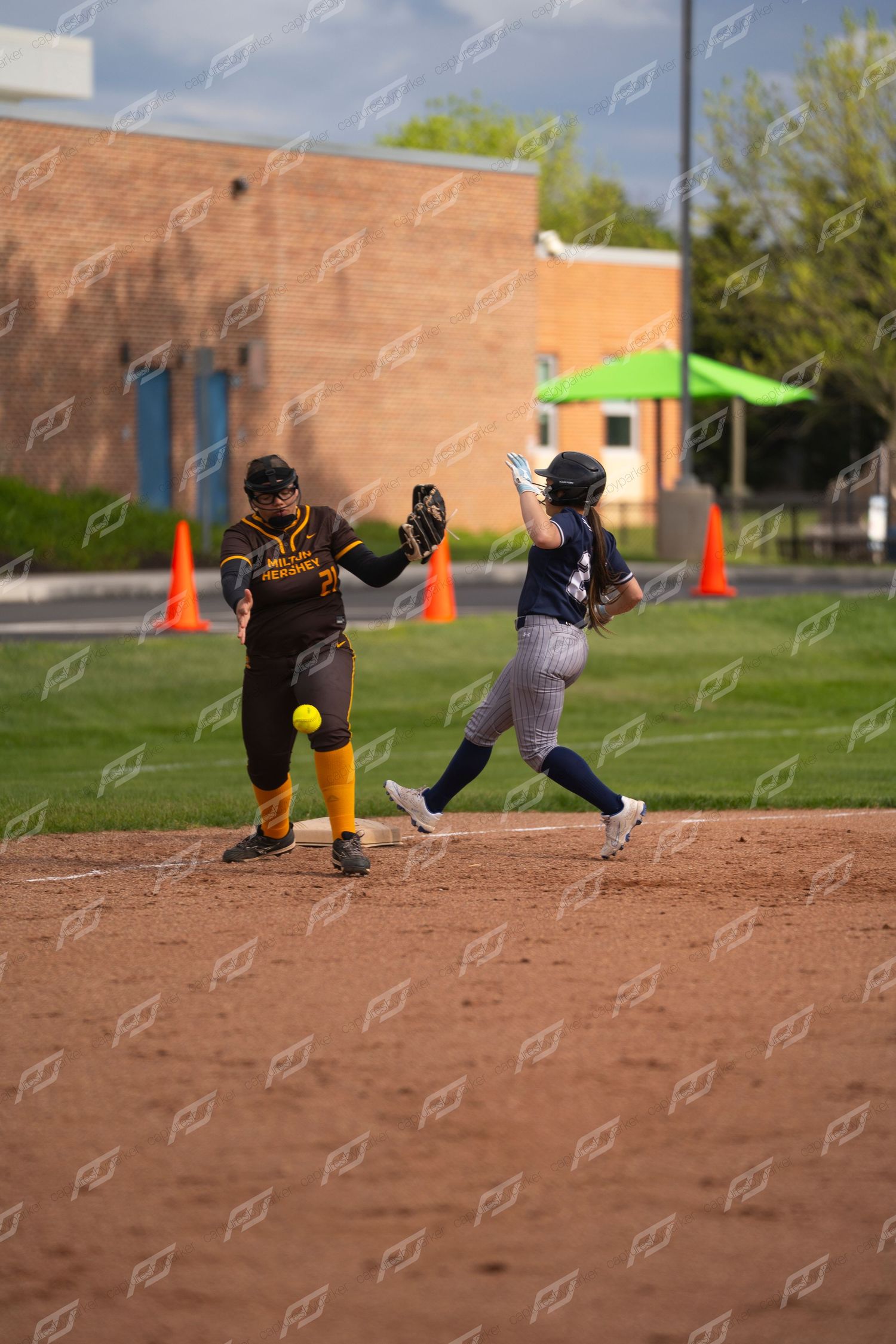 Navy-uniformed softball player races to first base as opposing first baseman catches the throw.