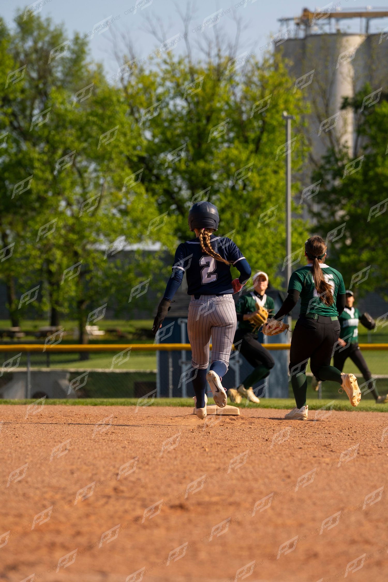 Softball player number 2 in navy uniform running bases as opposing green-uniformed fielder pursues play.