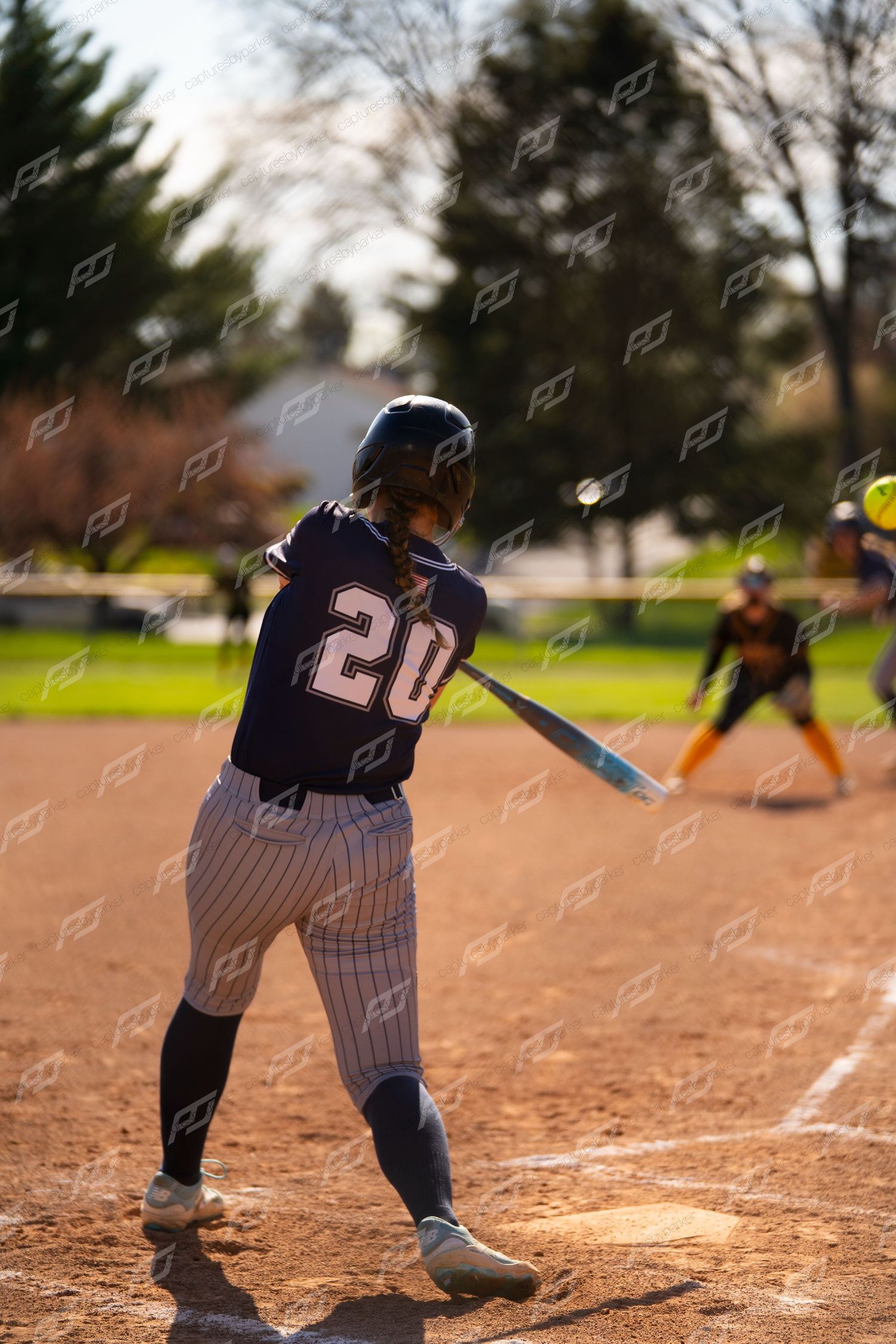 Softball player number 20 swings at incoming yellow pitch during sunny outdoor softball game.