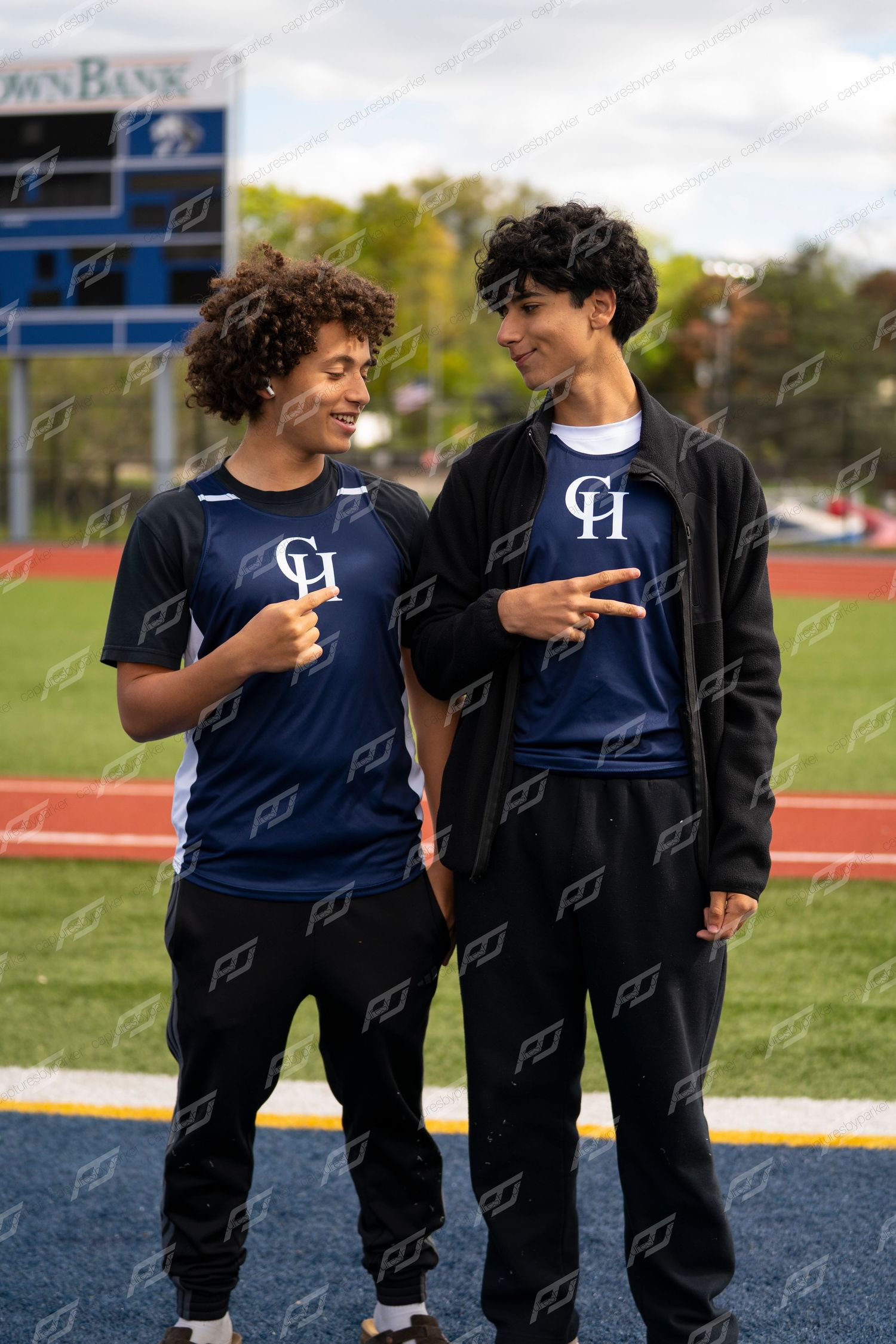 Two smiling male CH track athletes in navy uniforms laughing and pointing at each other on a sports field.