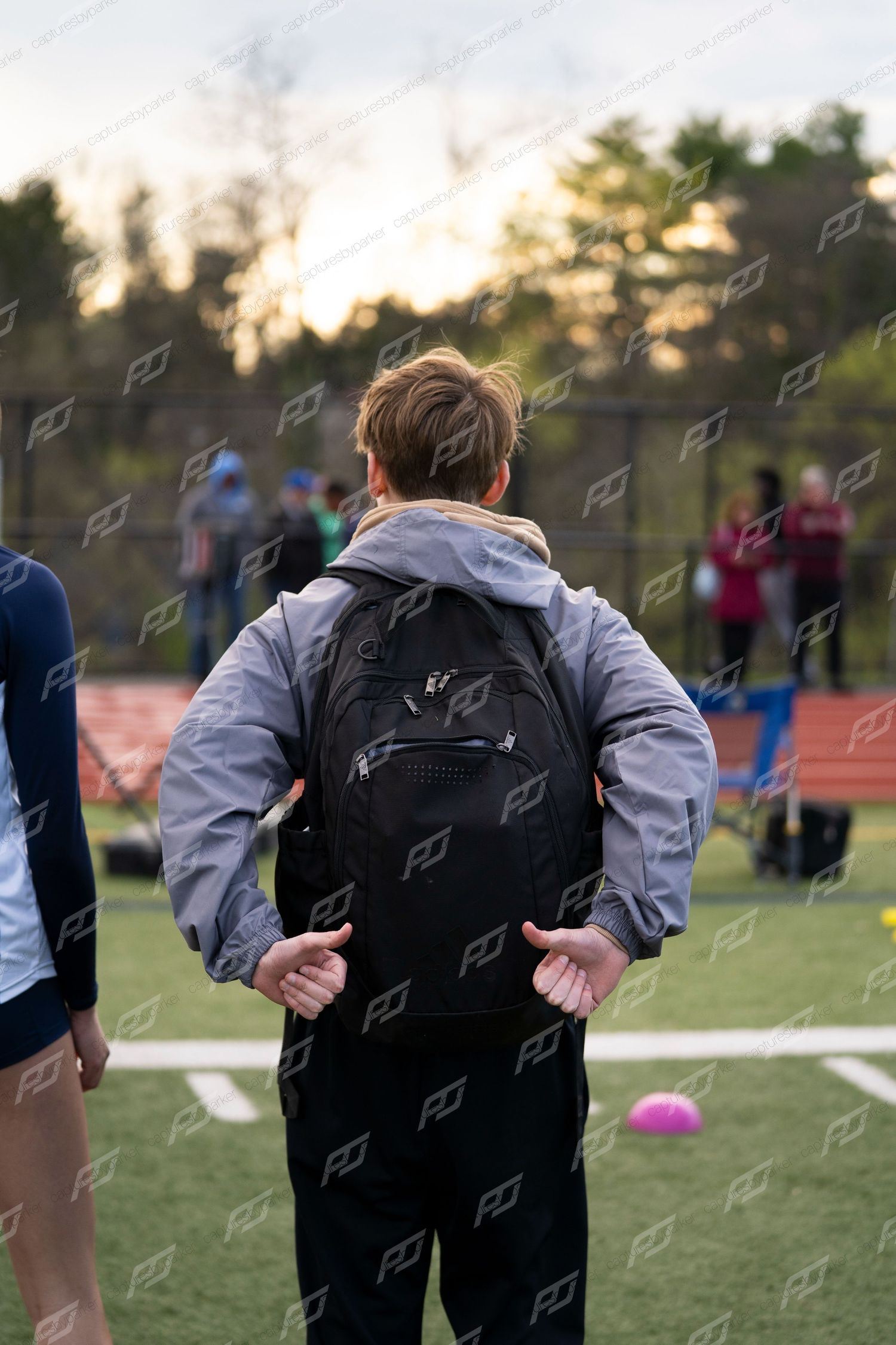 Male athlete with black backpack viewed from behind watches activity at outdoor track meet.