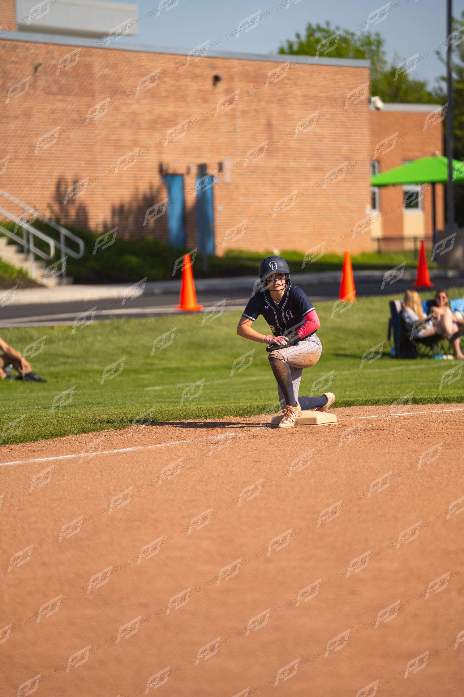 Softball player crouches in ready position on base during a sunny outdoor game.