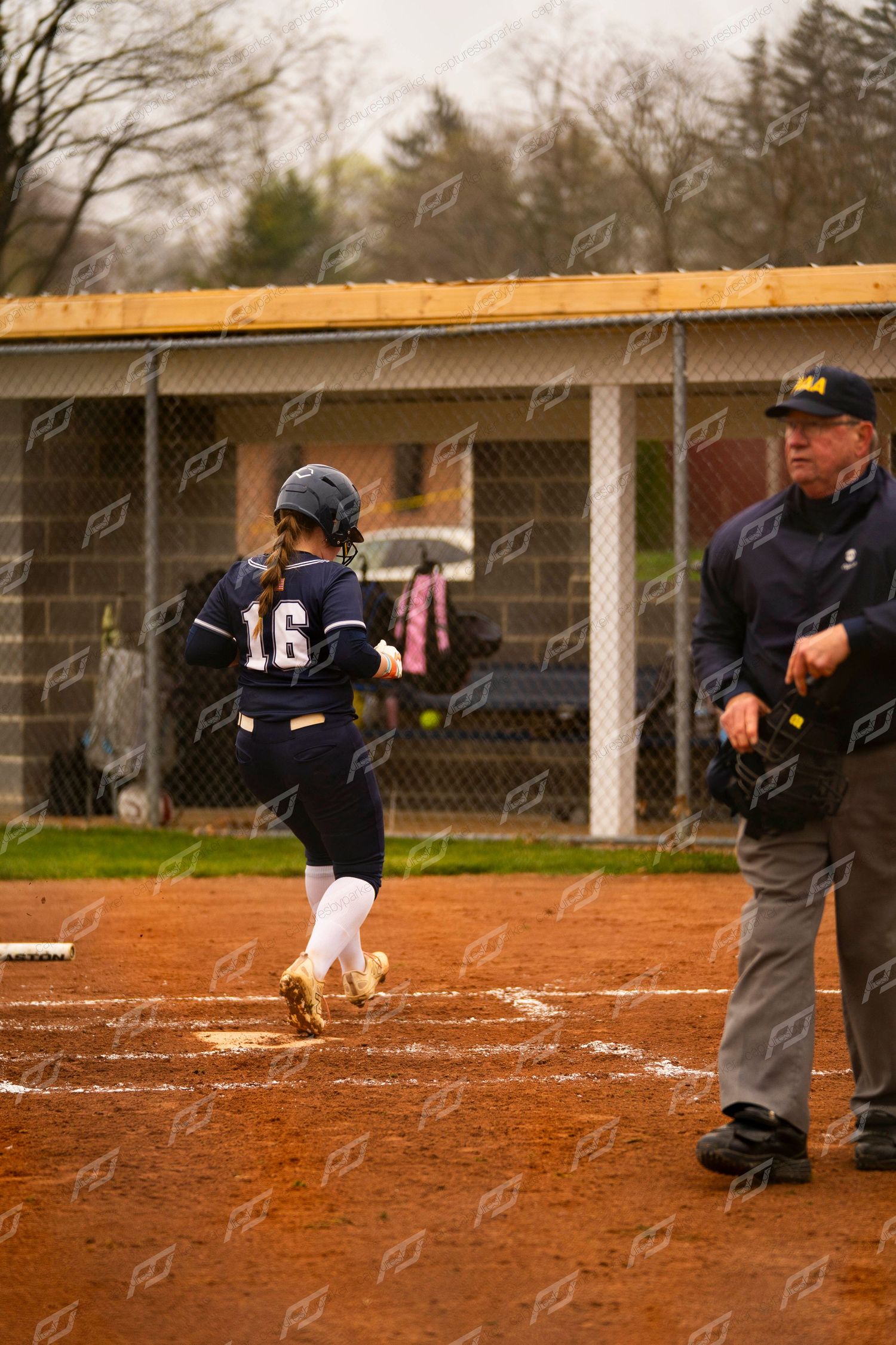 Female softball player #16 in navy uniform crosses home plate as umpire watches closely.