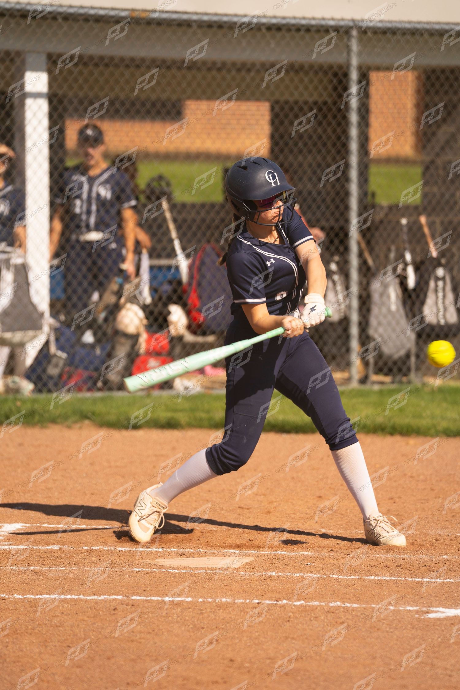 Female softball batter in navy uniform making contact with a yellow ball at home plate.