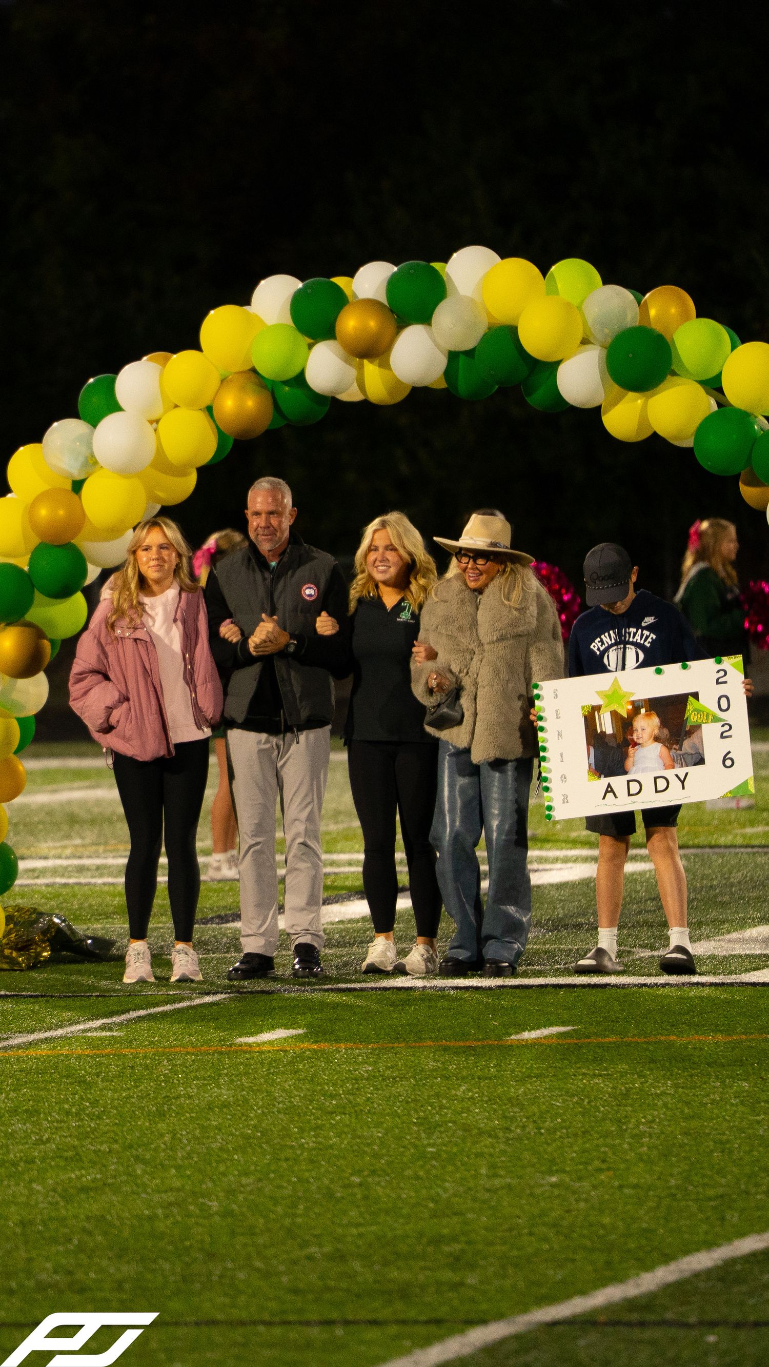 Group poses under green, gold, white balloon arch at senior night ceremony with Addy 2026 poster on football field.